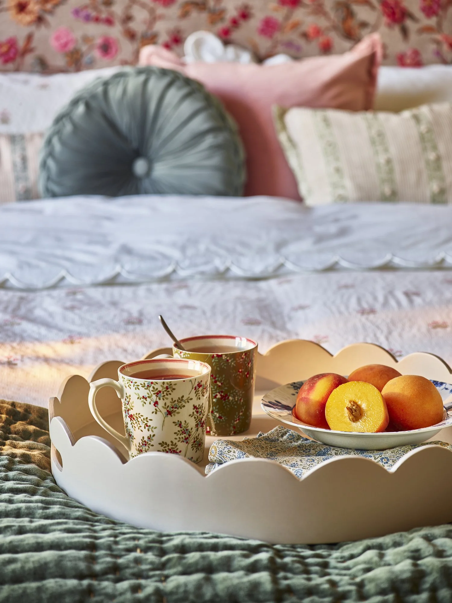 A tray with two mugs of hot beverage and a bowl of peaches on a bed, with bedsheets and pillows in the background.
