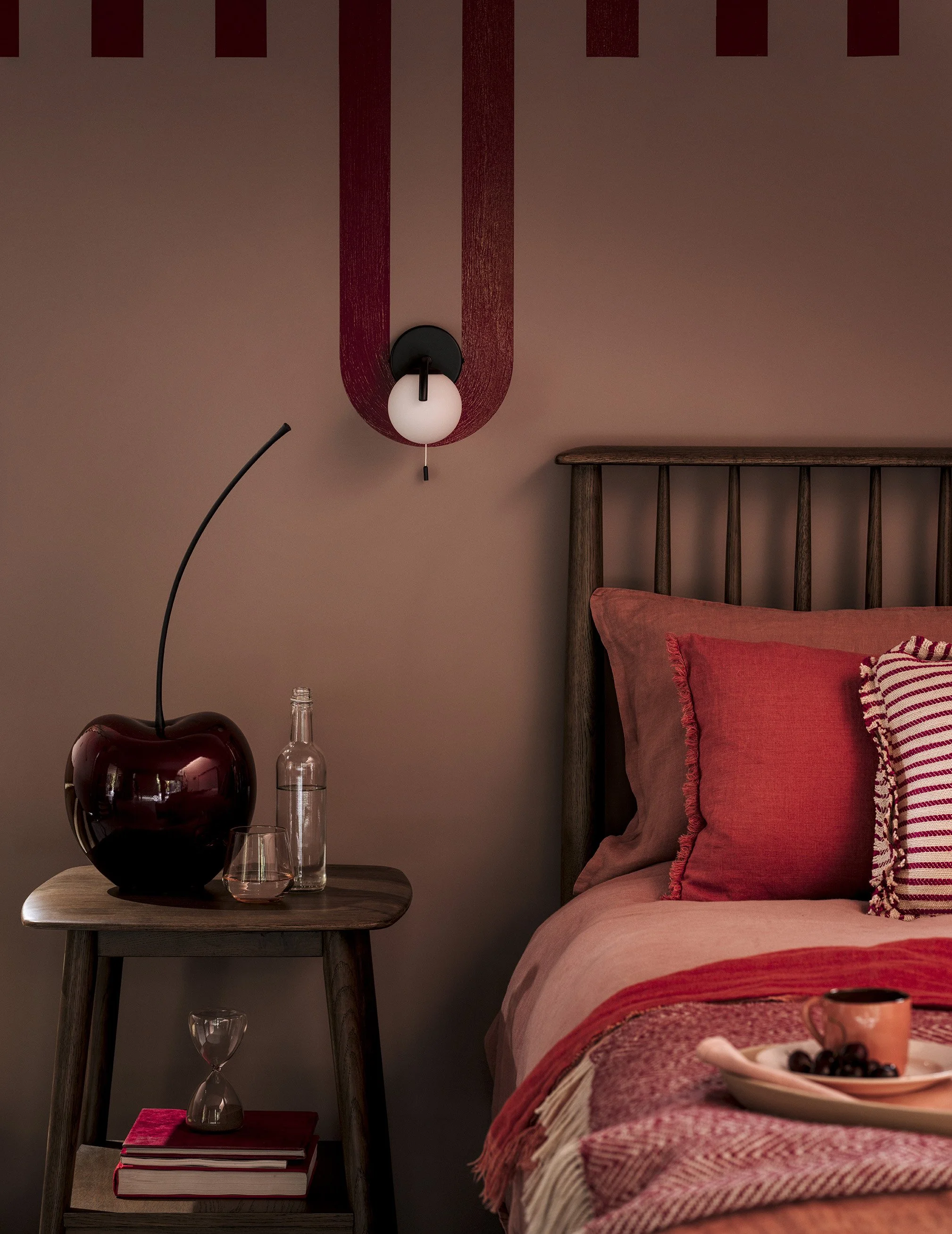A bedroom with a wooden bed frame, red and pink bedding, and pillows. A bedside table holds a cherry-shaped vase, a glass bottle, and a glass. A small table below has books and an hourglass. Wall decor includes a black and white wall lamp and a penda