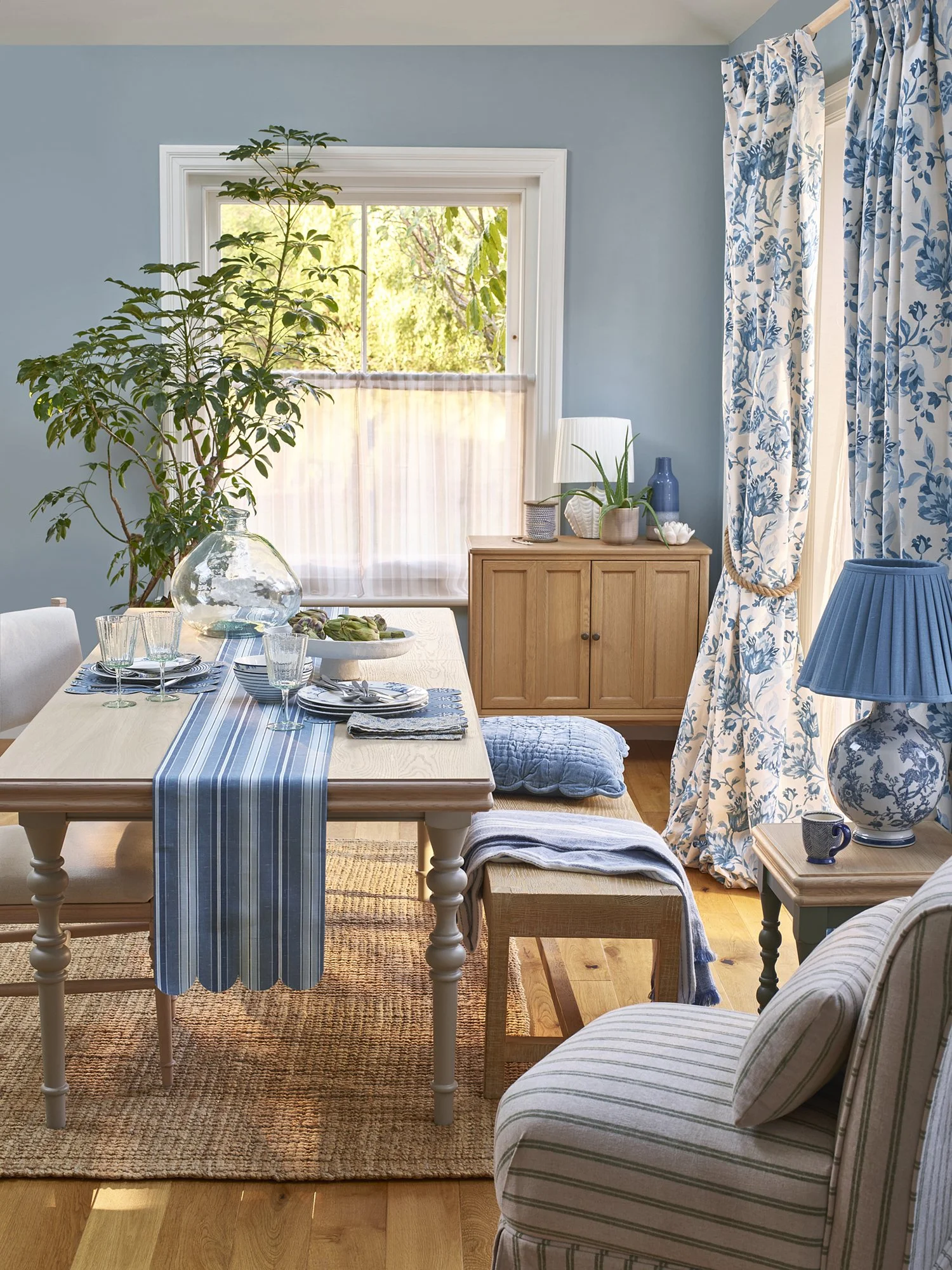 A cozy dining room with light blue walls, a wooden table decorated with blue-striped table runner and place settings, large window with white curtain, blue floral curtains, a sideboard with plants, lamps, and vases, and a striped armchair.