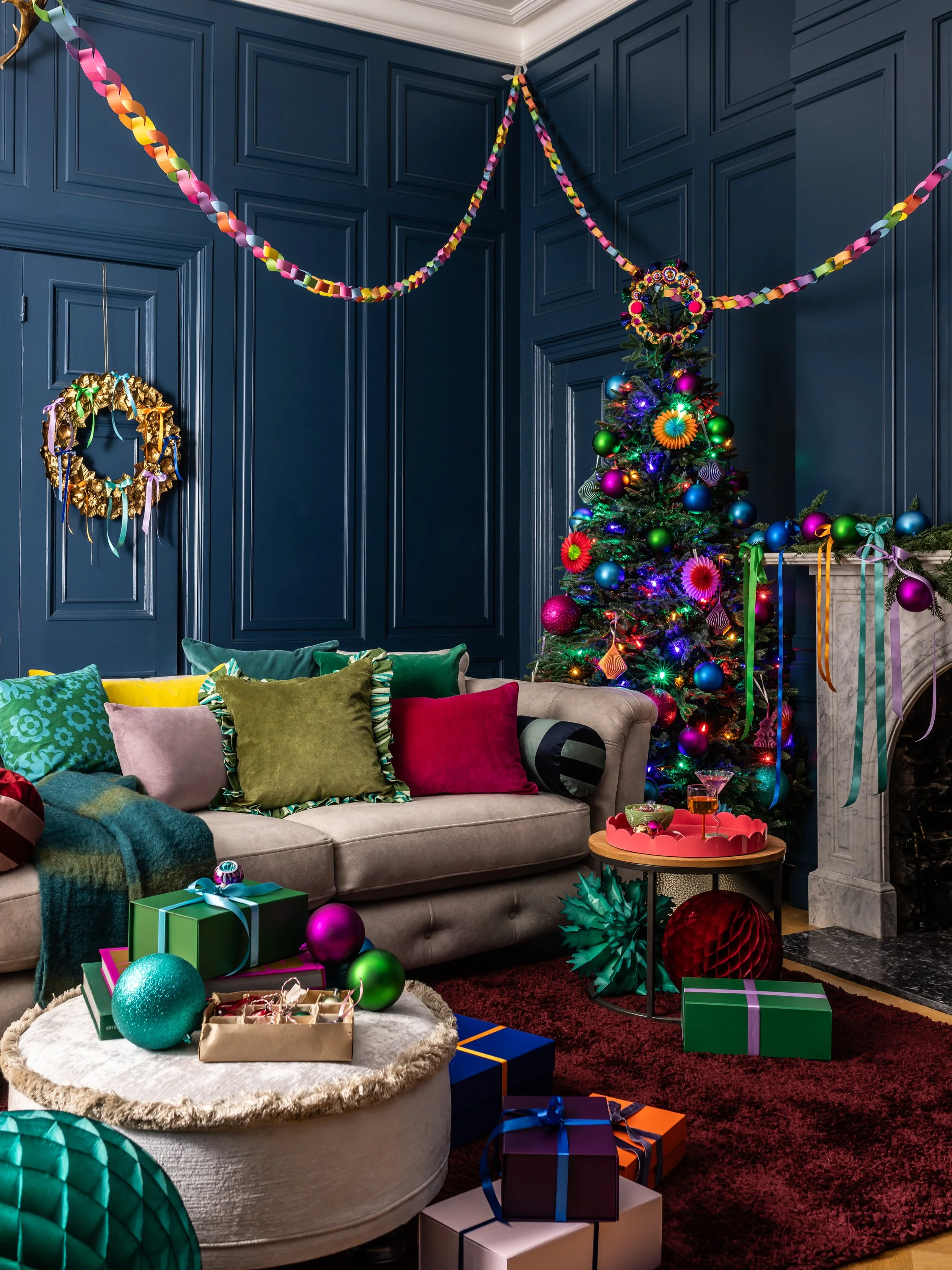 A decorated living room with a Christmas tree, colorful ornaments, wrapped presents, and festive decorations on dark blue paneled walls.