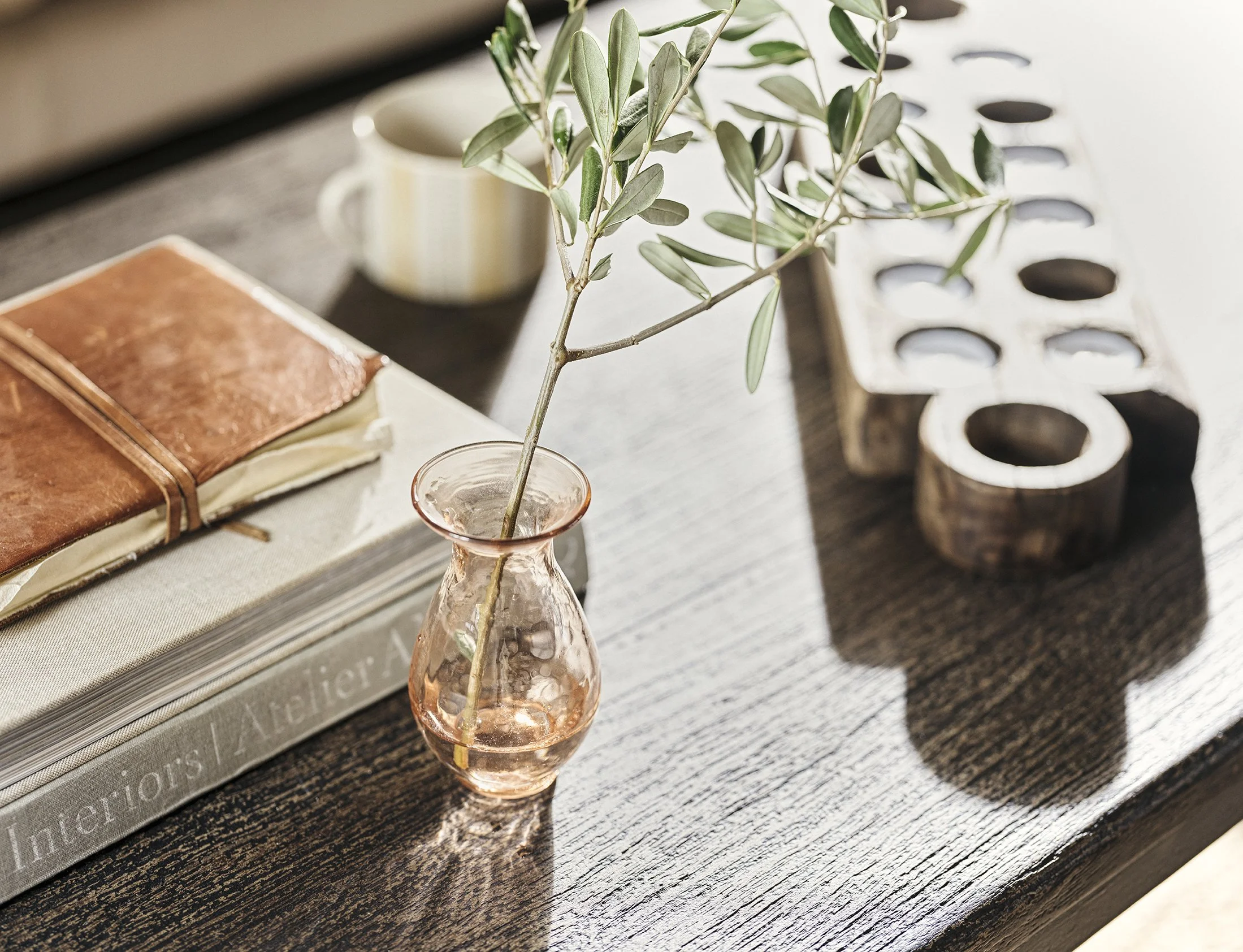 A small pink glass vase with a branch of green leaves, a brown leather journal, a beige book, a white coffee mug, a wooden alphabet block with the letter 'R', and a wooden block with circular cutouts on a dark wooden surface, with sunlight casting sh