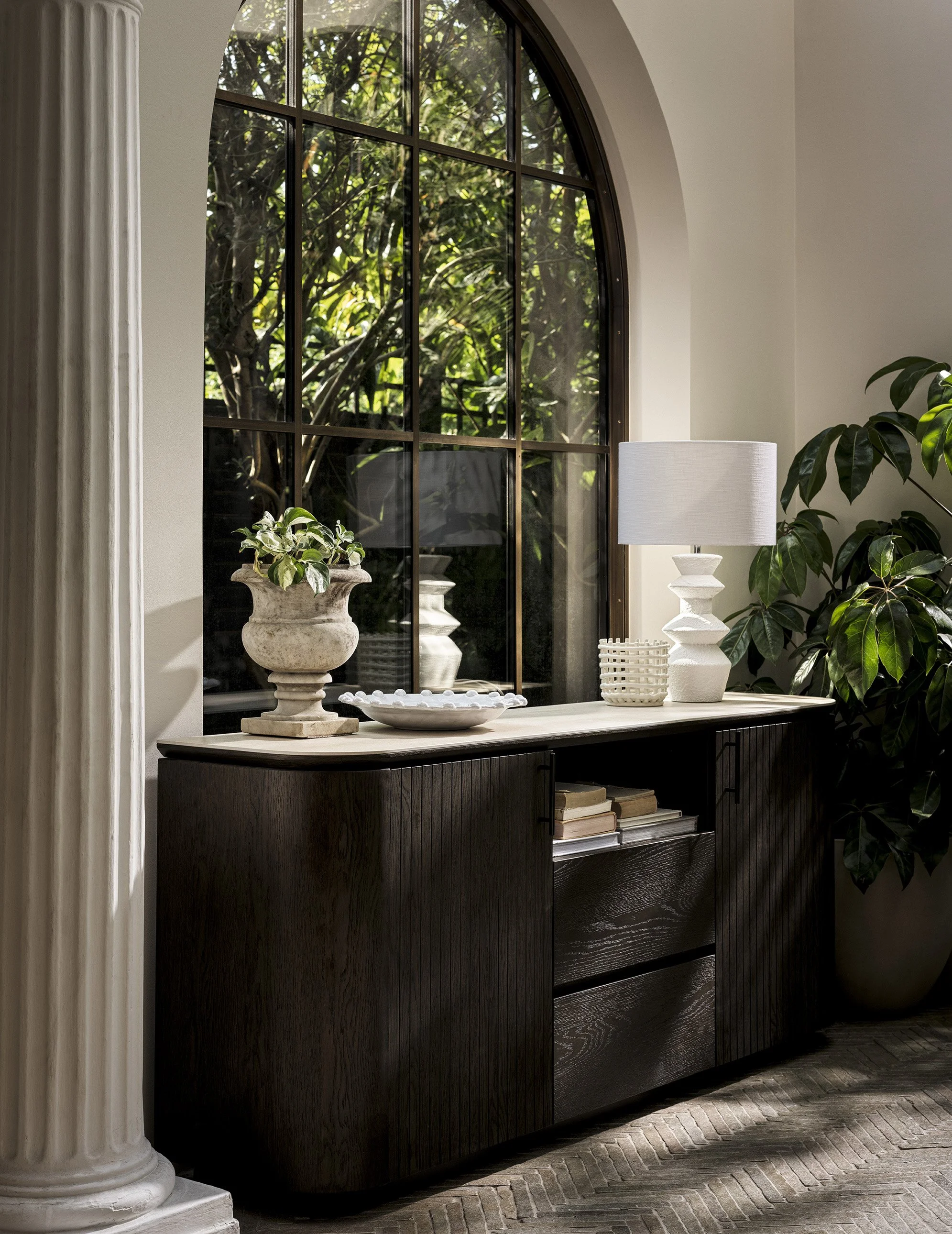 Interior view of a sideboard with decorative items, including a potted plant in a classical-style urn, a large white table lamp, a woven basket, and a white decorative dish, placed in front of a large arched window with black framing, overlooking gre