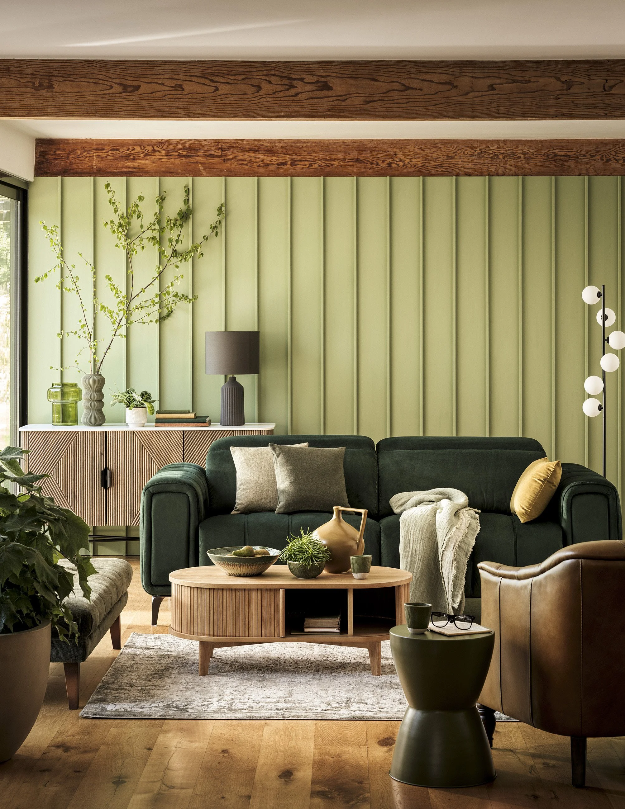 Living room with green wall, wooden beams, green sofa, brown leather chair, side table, decorative items, and plants.