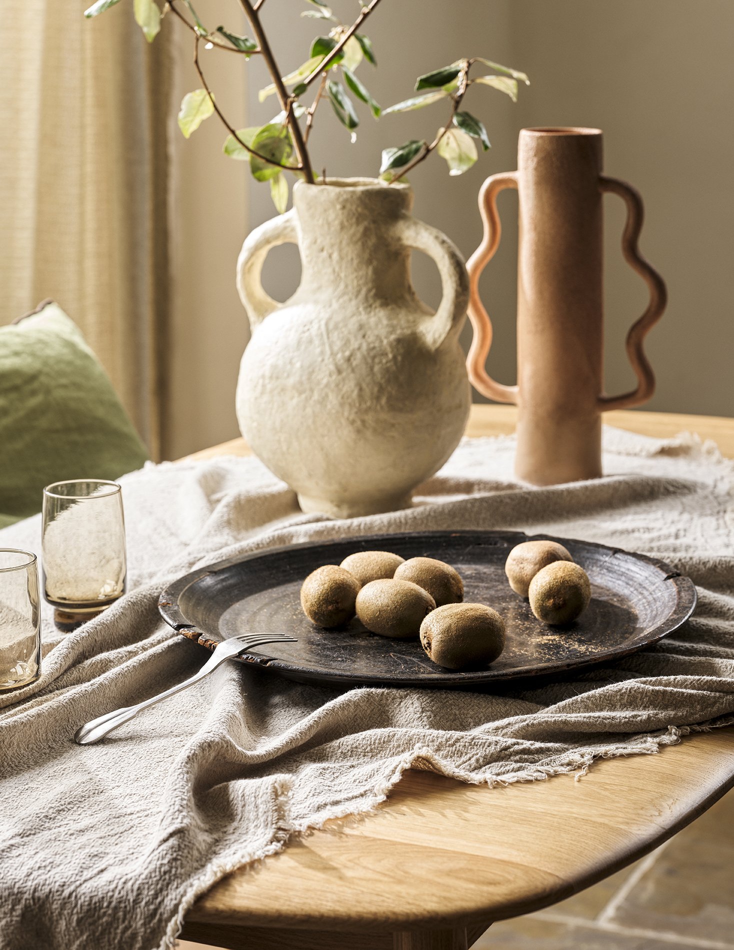 A wooden dining table with a beige cloth runner, featuring a black round tray with round brown fruits, a fork, and two empty glasses. In the background, there is a textured white ceramic vase with green plant leaves and a tall, wavy-collared, pinkish