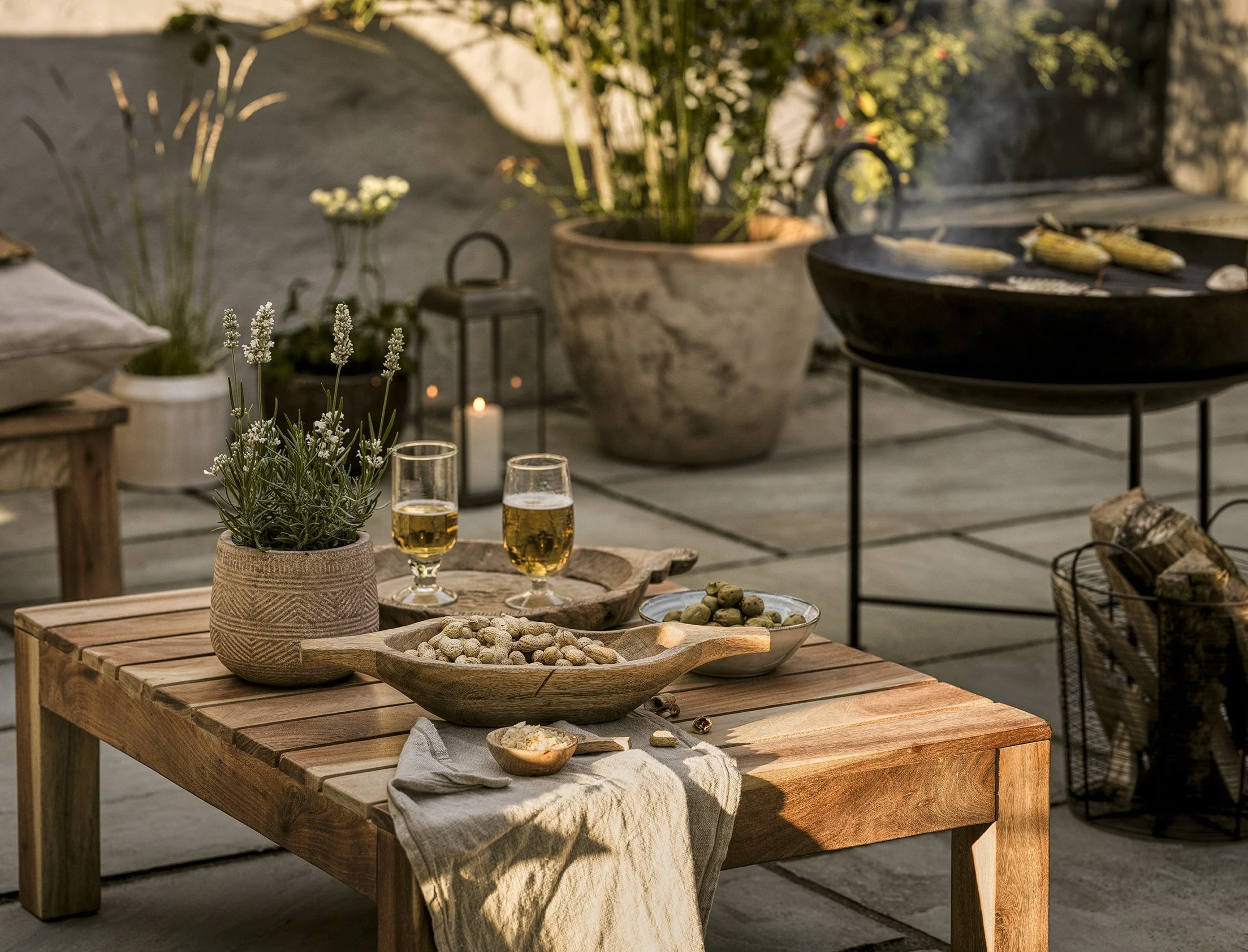 An outdoor patio table with snacks, drinks, and plants, including a potted lavender, two glasses of beer, and bowls of nuts and olives. In the background, there is a large pot with green plants, a candle, lantern, and a grill with corn on the cob coo