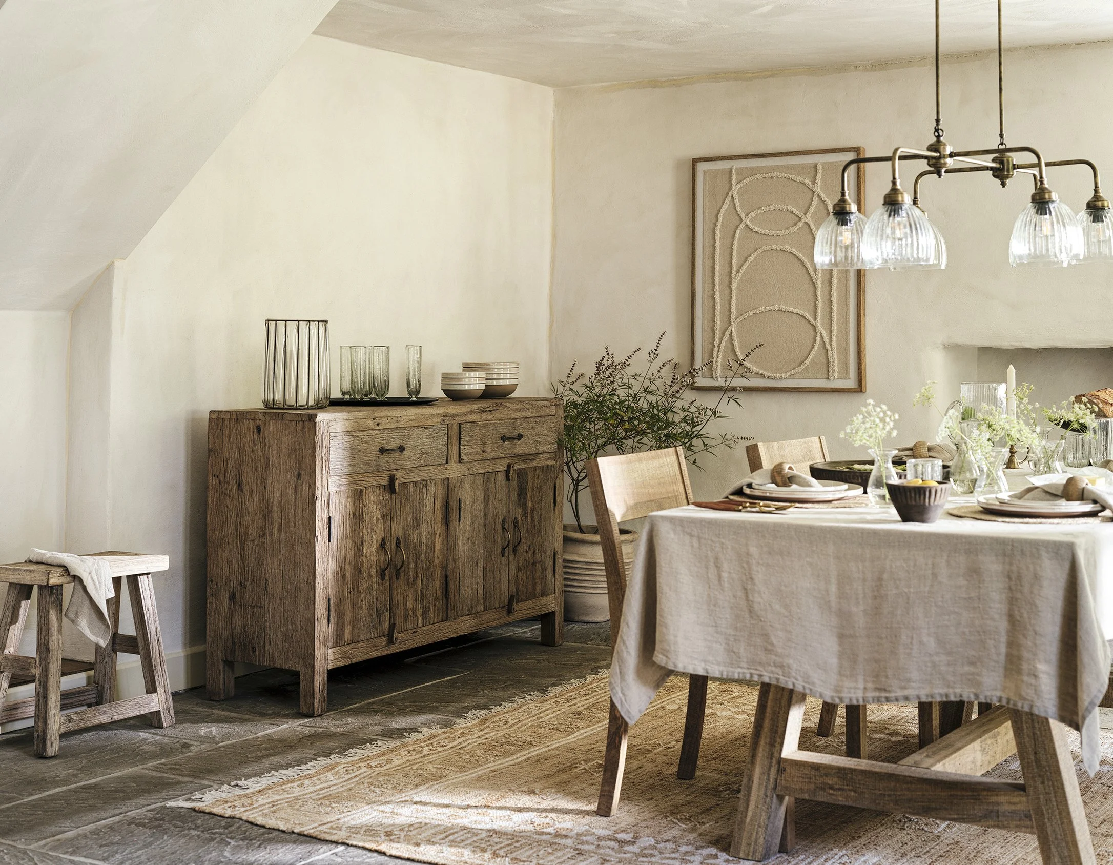 A rustic dining room with a wooden sideboard, a table set with plates, bowls, and glasses, and a chandelier hanging above. The room has a light, neutral color palette with decorative art on the wall.