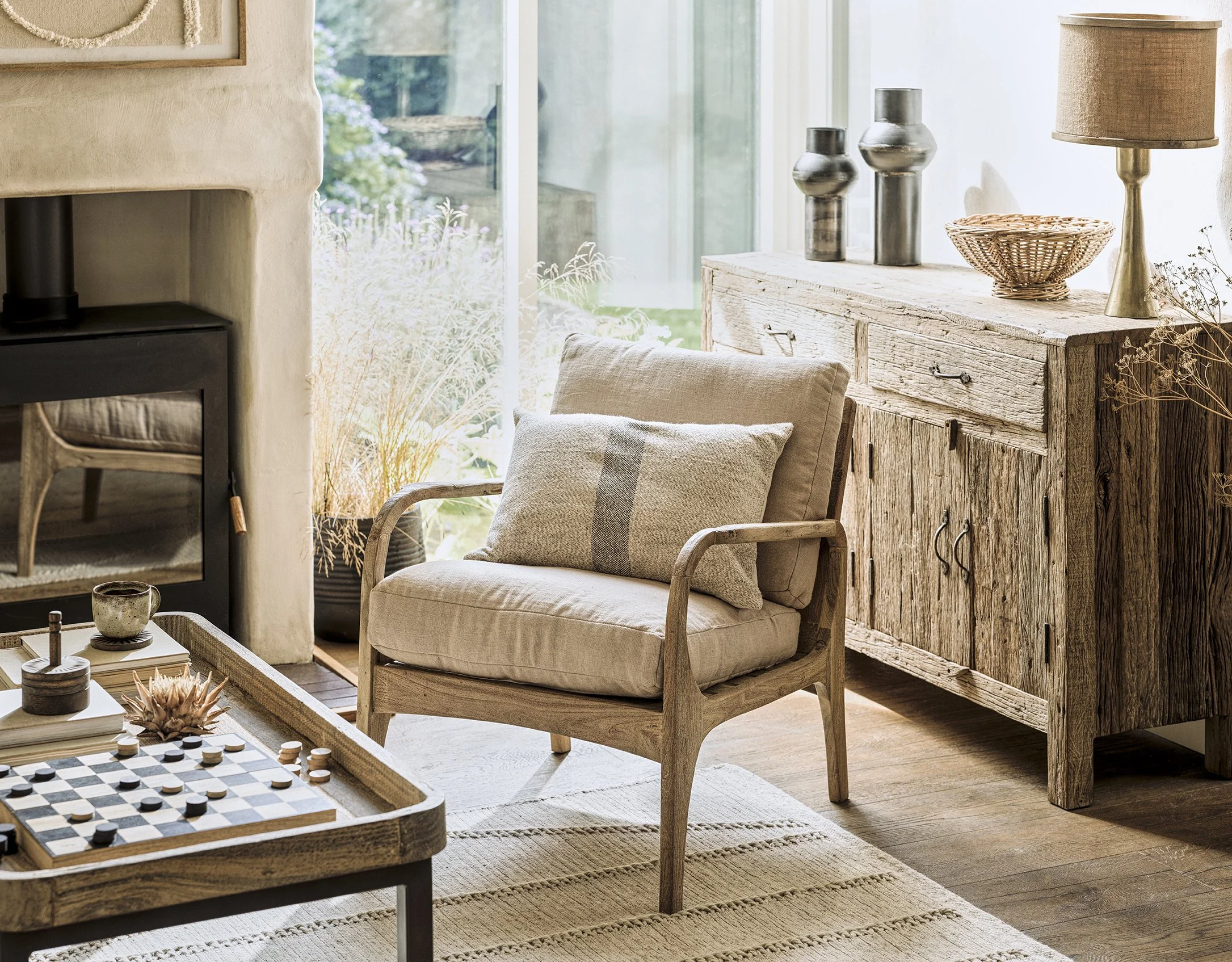 Cozy living room with a beige armchair, a wooden sideboard with decorative vases and a lamp, a fireplace, large window with natural light, and a coffee table with board games and cups.