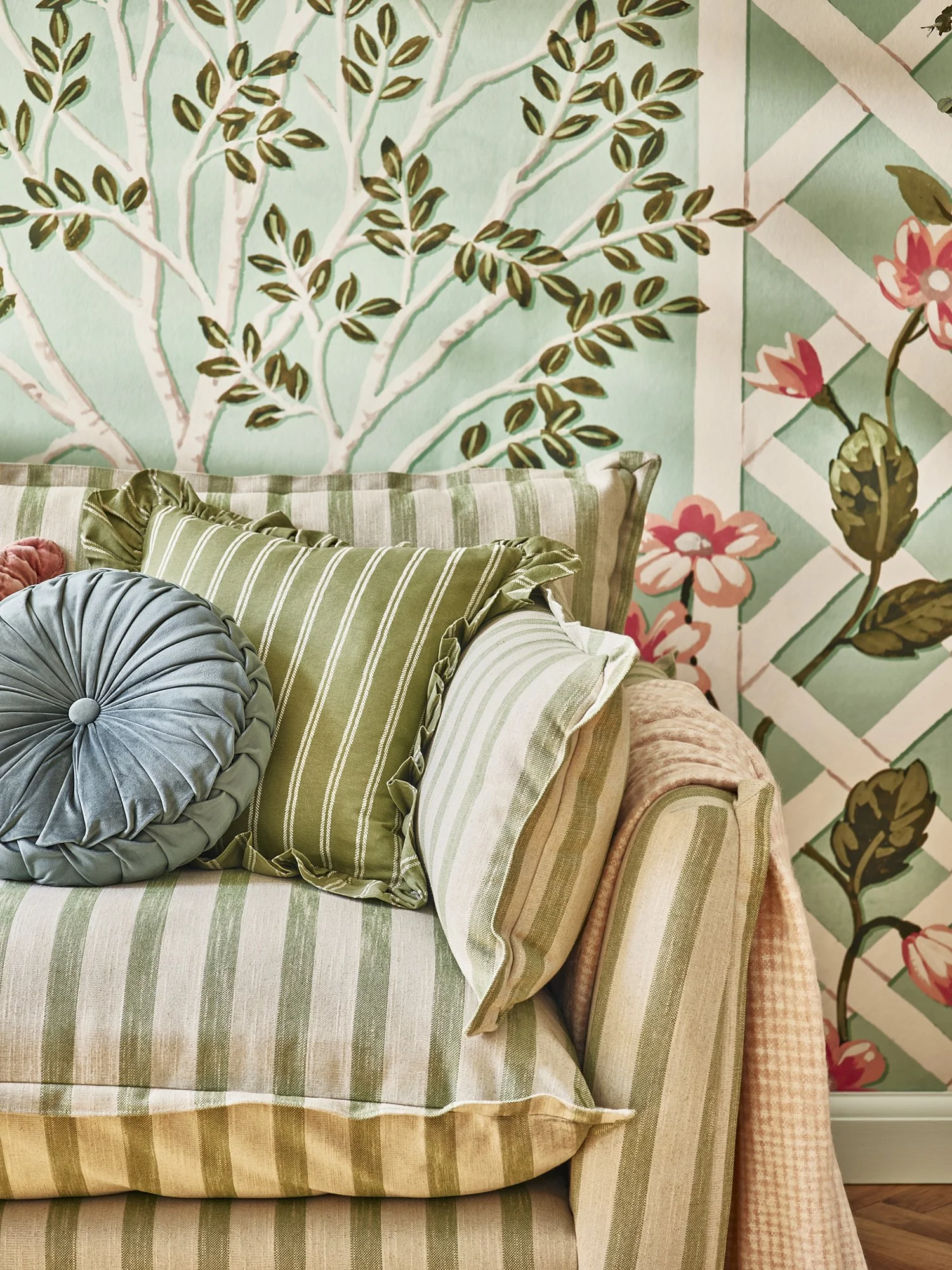 Close-up of a sofa with striped green and beige upholstery, decorated with green and striped pillows, set against a floral wallpaper with trees, leaves, and pink flowers.