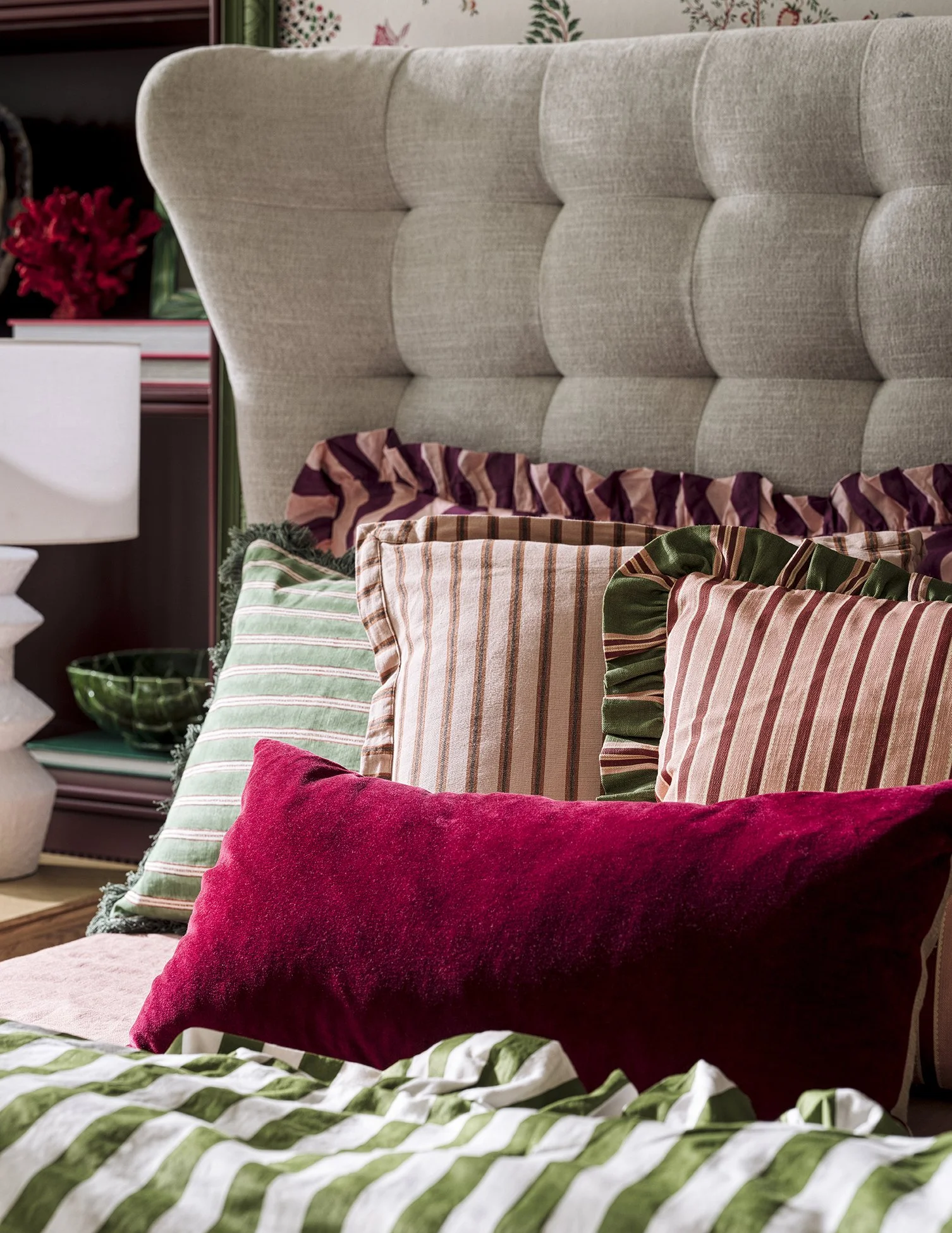 Close-up of a bed with multiple decorative pillows in various colors and patterns, including red, green, striped, and ruffled designs, against a beige tufted headboard.