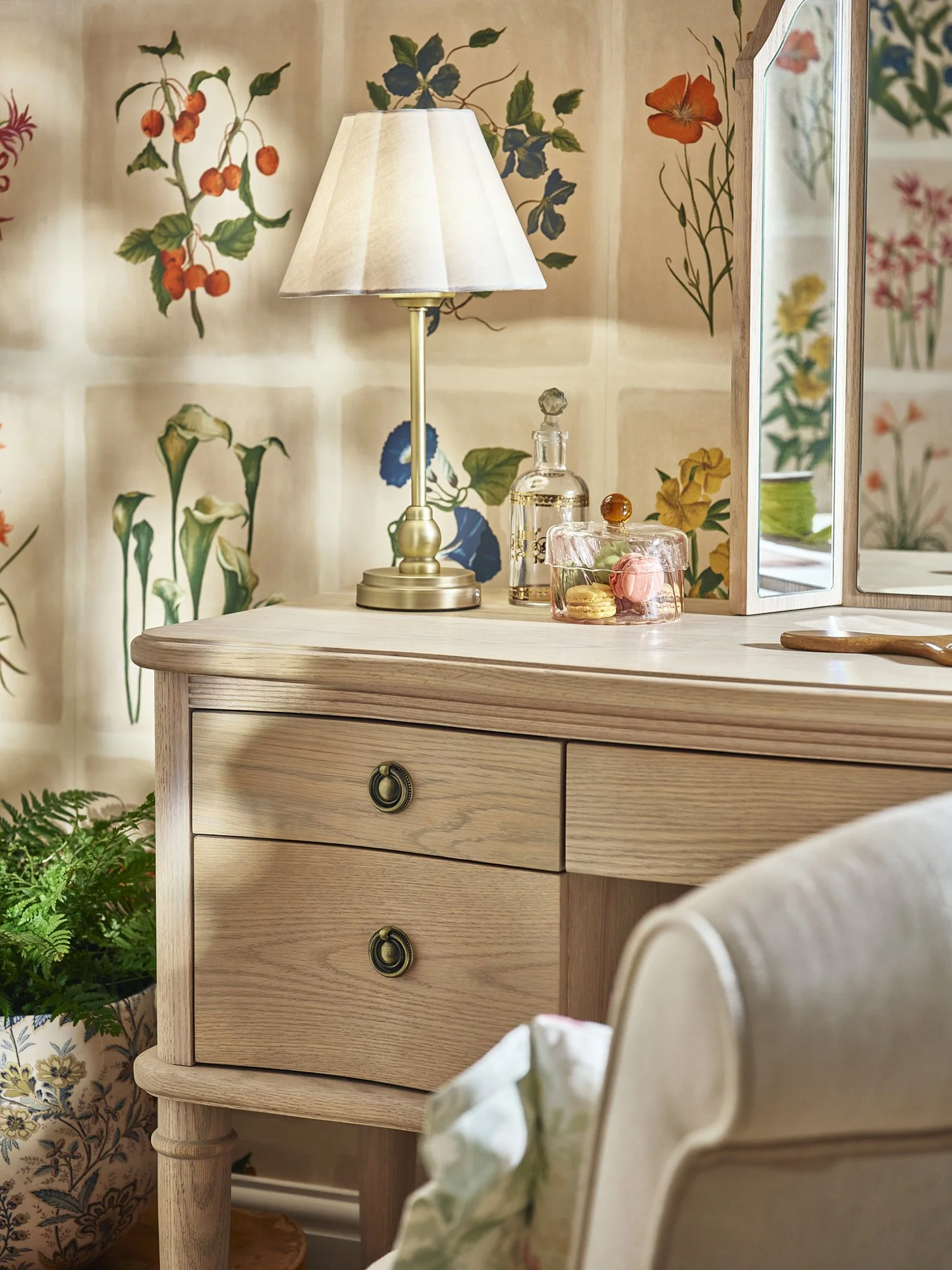 A wooden dresser with a brass table lamp, a glass decanter, a pink glass container with macarons, and a mirror on top. A botanical wallpaper with various flowers and a potted plant in a decorated ceramic pot are visible in the background.