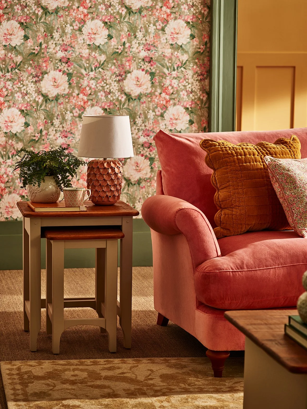 Living room corner with pink velvet sofa, yellow velvet pillow, floral wallpaper, wooden side table with a lamp, plant, and mug, beige rug, and green trim.