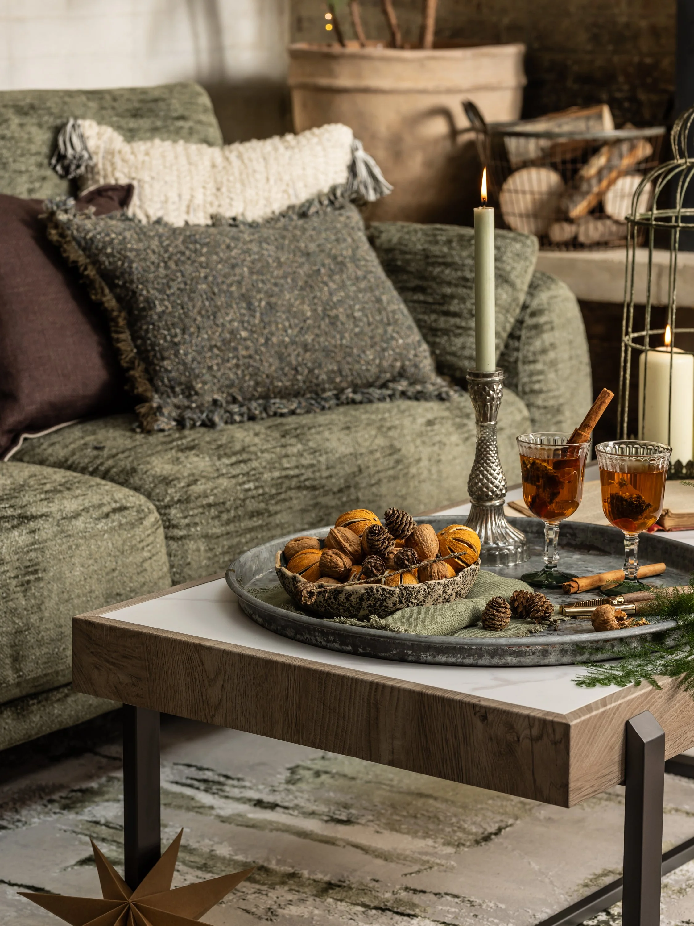 Decorative living room scene with a green couch, textured pillows, a wooden coffee table with pinecones, nuts, candles, and glasses of drink, in cozy warm lighting.