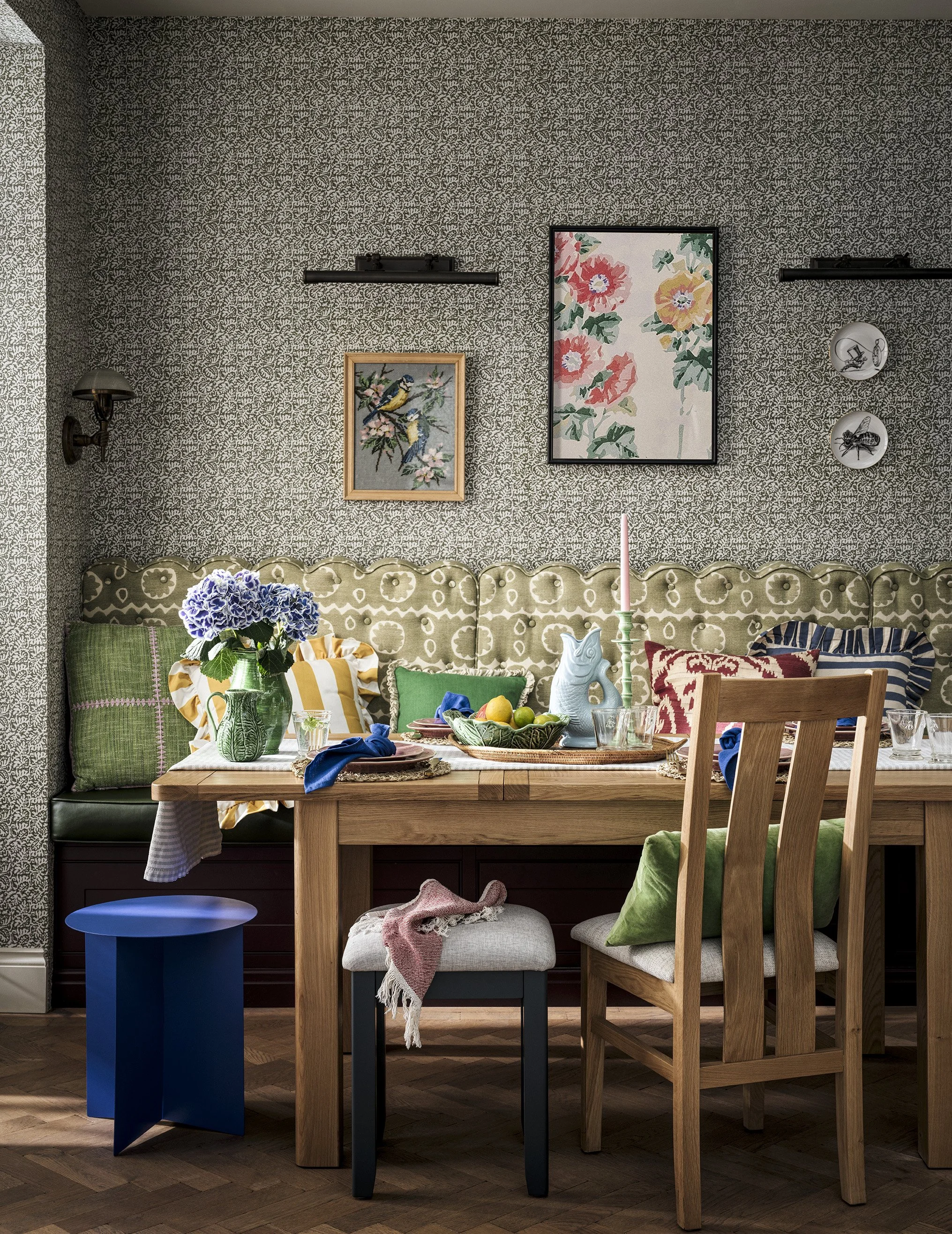 A cozy dining room with a wooden table decorated with a vase of blue flowers, fruit bowl, candelabra, and various pillows on a built-in green patterned banquette. The dining chairs include a wooden one with a green pillow and a small upholstered stoo