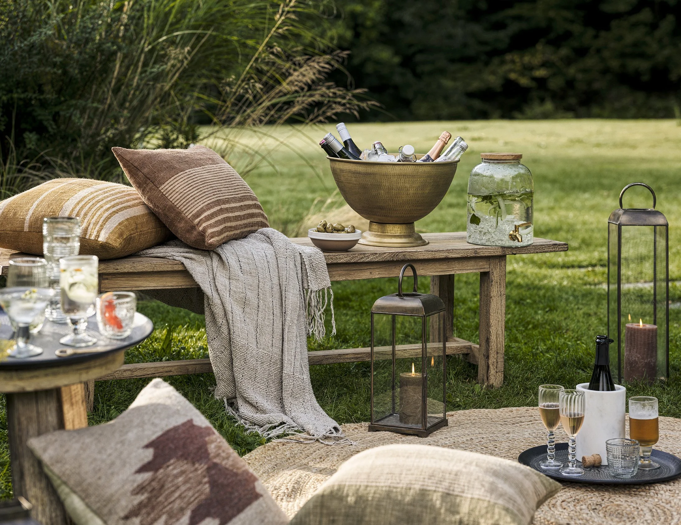 Outdoor picnic setup on a grassy area with cushions, a wooden table holding a bowl of bottles, candles, and drinks, and additional candles and glasses on a tray nearby.