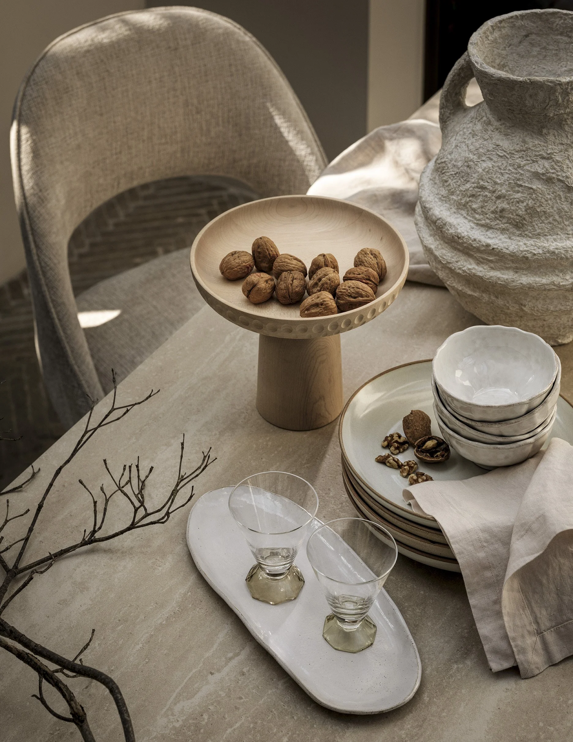 A dining table decorated with a wooden pedestal bowl filled with walnuts, a stack of white ceramic bowls, a white ceramic platter with walnuts and a linen cloth, a tray with two empty glasses, a large textured vase, some branches, and a stack of plat