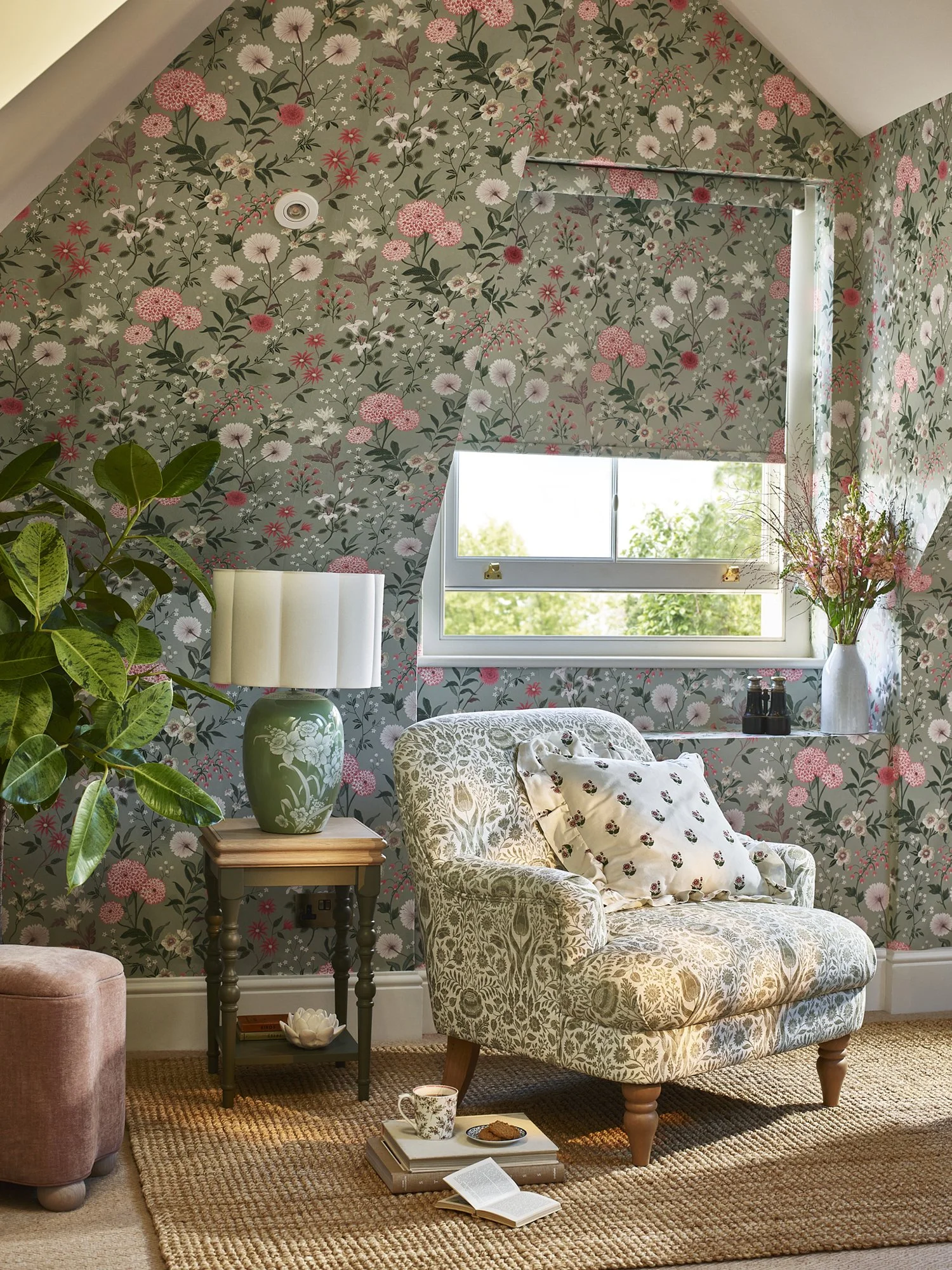 A cozy living room corner with floral-patterned wallpaper, a window with a roller blind, a green-and-white lamp on a wooden side table, an upholstered armchair with patterned fabric and pillows, a potted plant, a vase with flowers on the window sill,