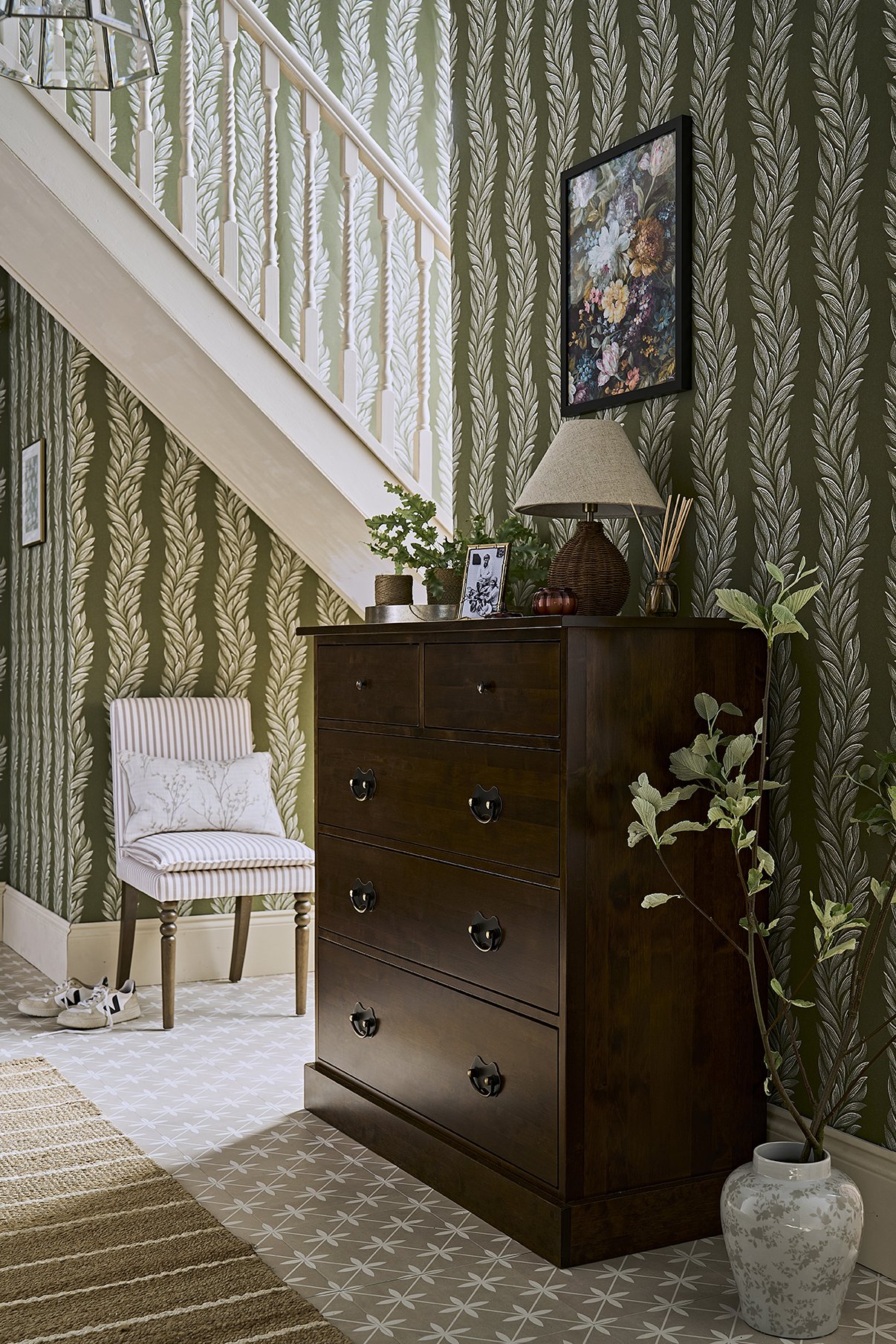 A cozy entryway corner with a dark wood dresser, a small upholstered chair with striped fabric and a pillow, potted plants, a table lamp, and framed photos. The wall has green leaf-patterned wallpaper, and the floor has geometric patterned tiles.