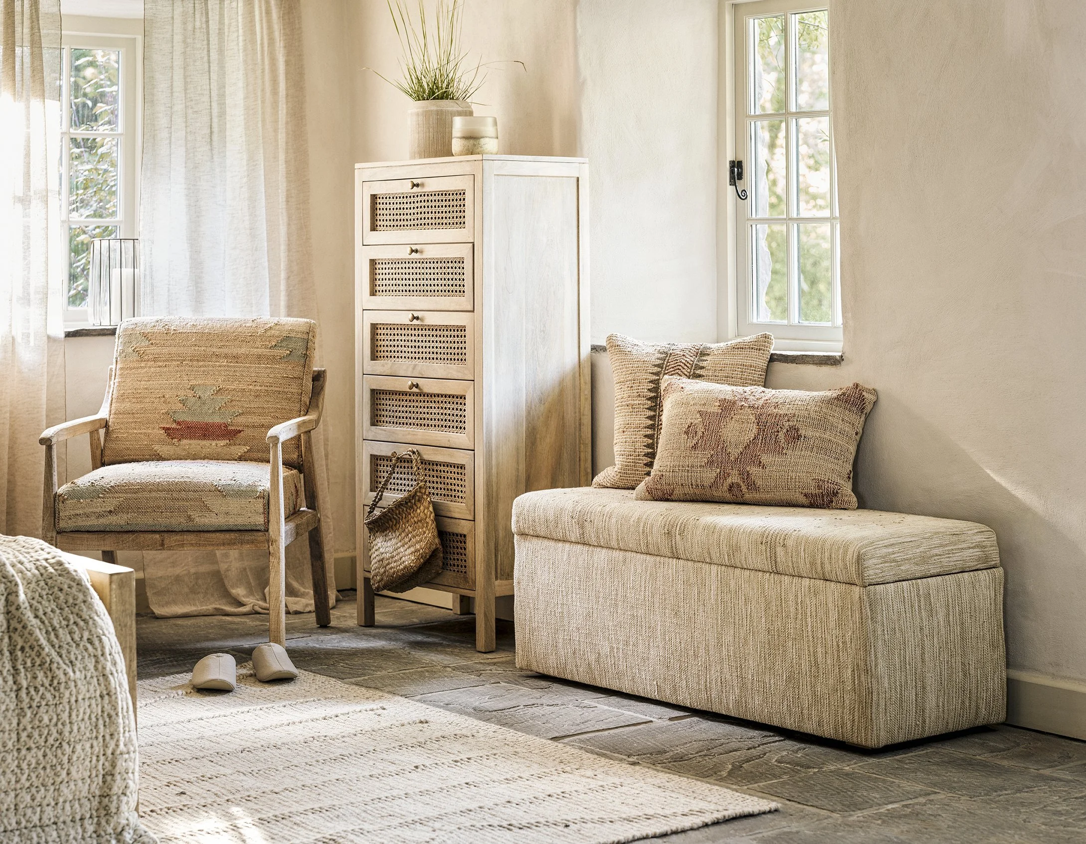 A cozy, well-lit room featuring a wooden armchair with a woven cushion, a tall white dresser with woven drawer fronts, a cushioned bench with two pillows, and two windows with sheer curtains allowing sunlight to brighten the space.