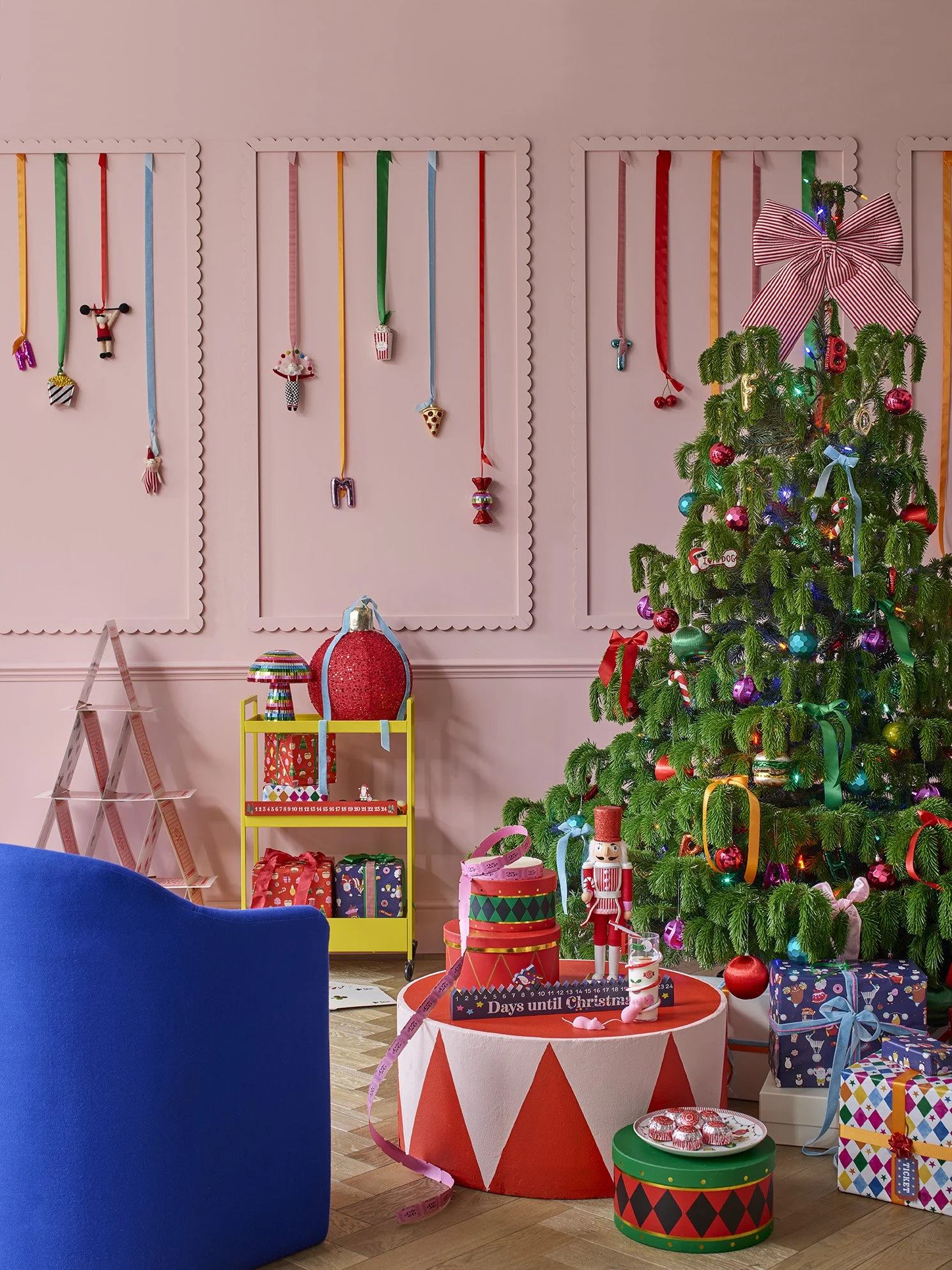 Decorated Christmas tree with ornaments and lights in a festive room with pink walls, surrounded by wrapped gifts and holiday decorations.