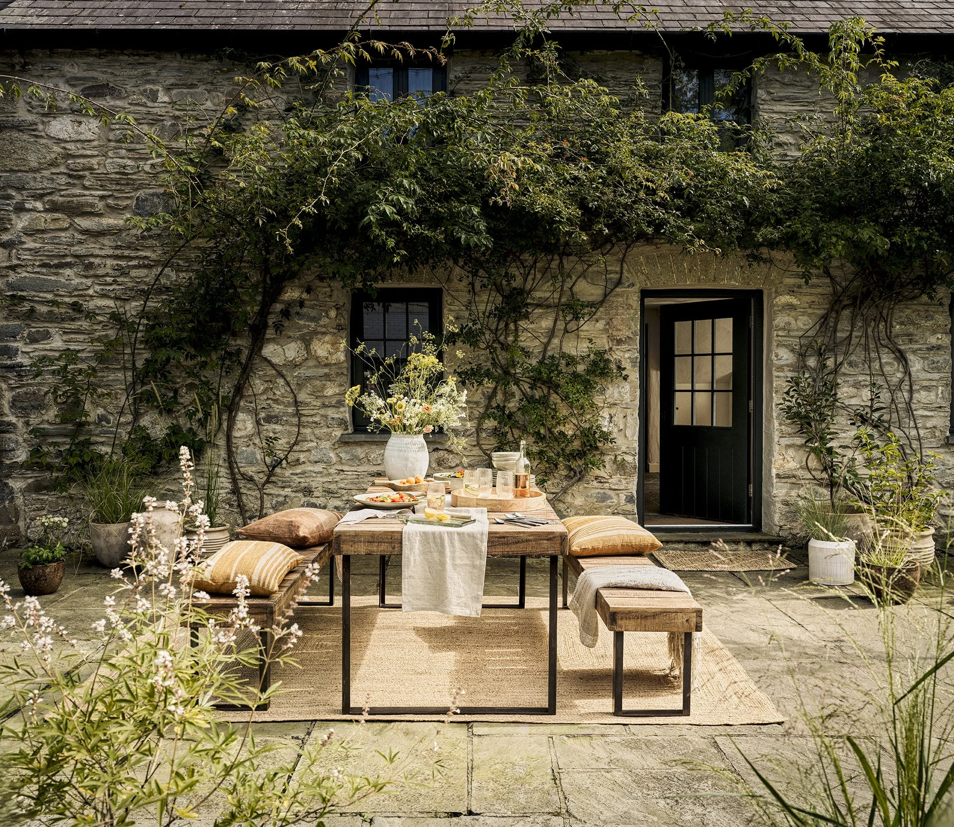 Outdoor dining setup with a rustic wooden table, several pillows on benches, a large vase with flowers, and a stone building with vines in the background.