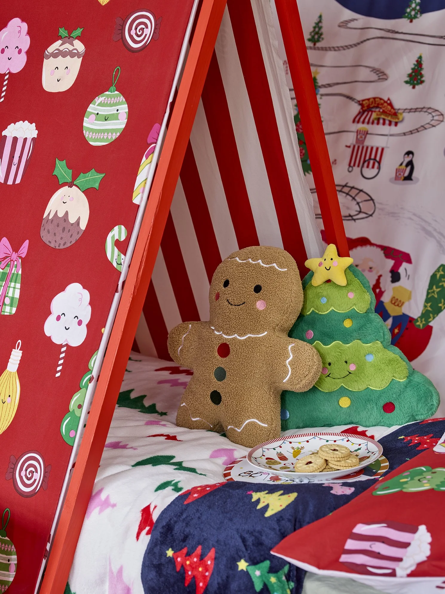 Decorative Christmas scene with a gingerbread man plush toy, a plush Christmas tree, a plate of cookies, and festive fabric and wallpaper with holiday designs.