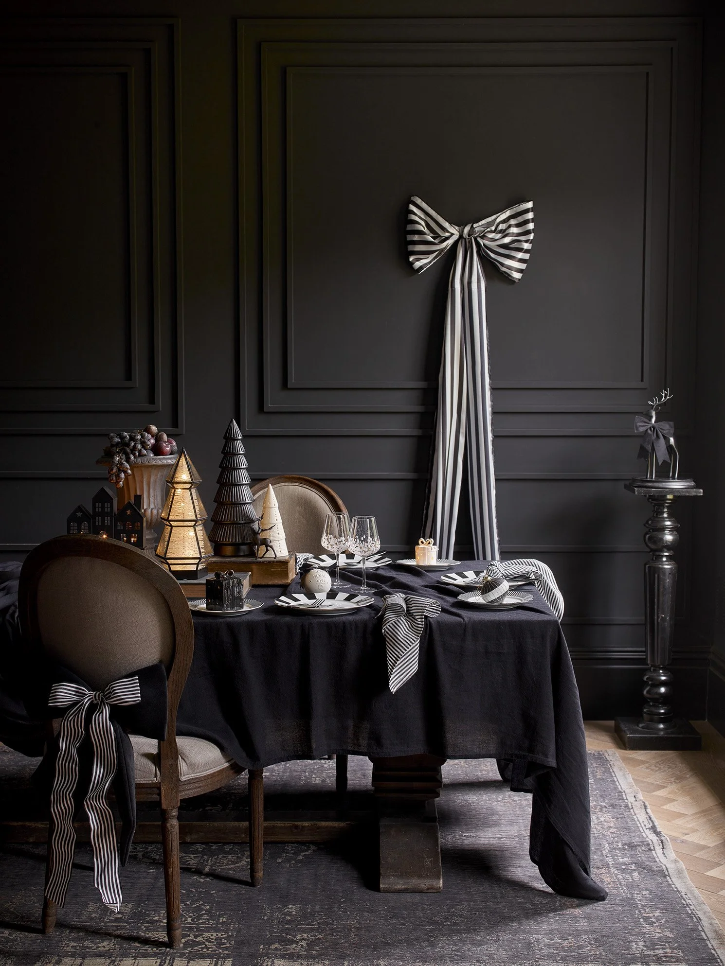 A dark-themed dining room with a black tablecloth on a round table, decorated with striped black and white ribbons on chairs, and holiday decorations including cone-shaped trees and a reindeer figurine with a bow, set for a meal with glasses and plat