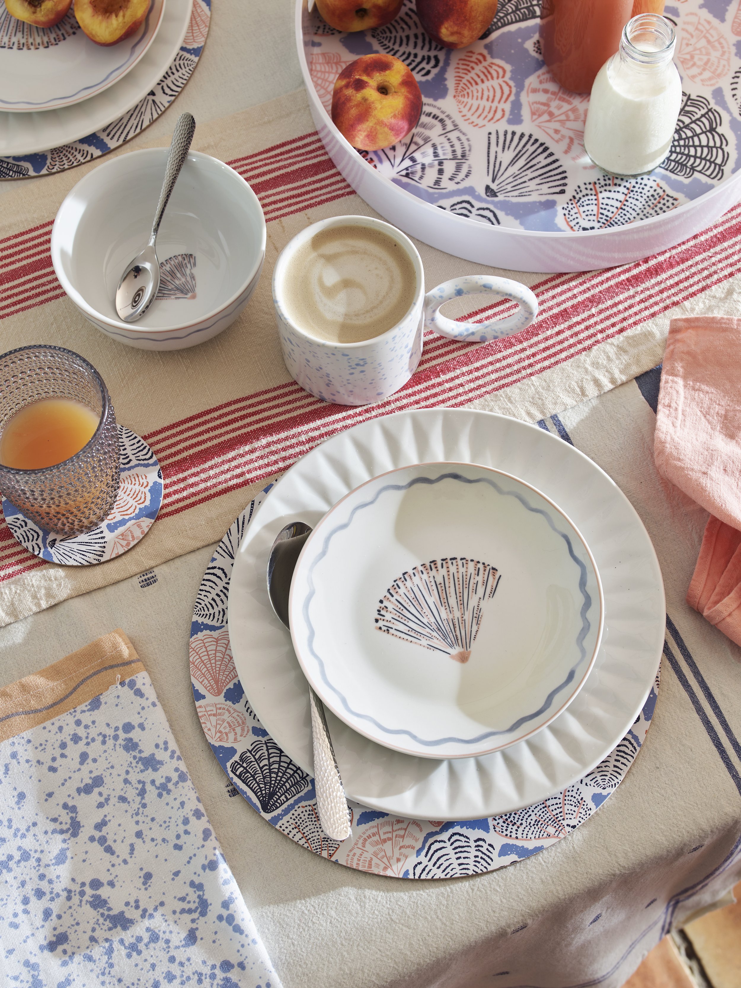 A table setting with patterned plates, bowls, and cups, including peaches, a glass of orange juice, a cup of coffee, a small bottle of milk, and a pink cloth napkin on a table with a decorative tablecloth.