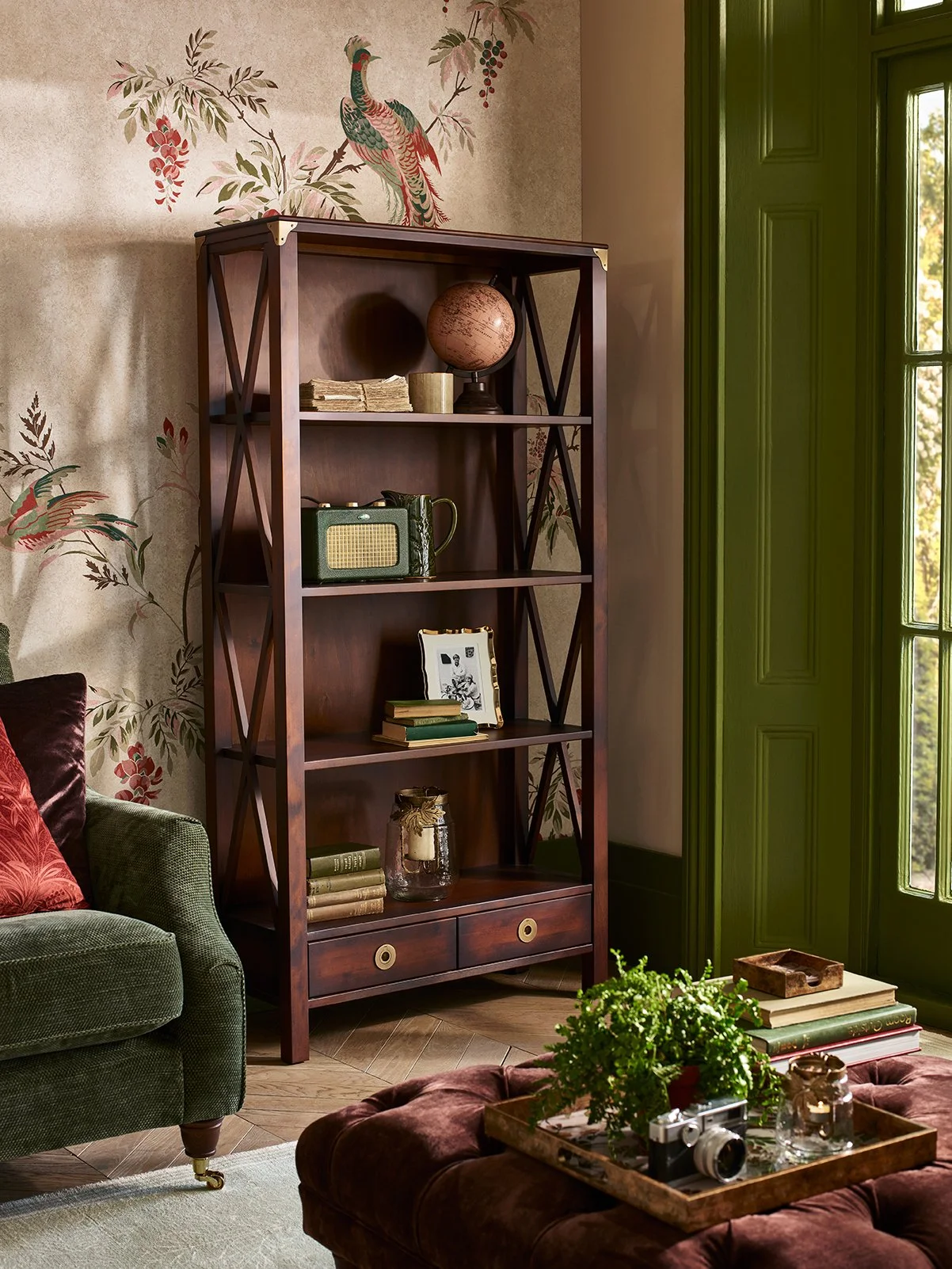 A living room corner with a wooden bookshelf against a decorative wallpapered wall showing a peacock and a tree. Part of a green armchair with a red pillow and a tufted ottoman with a tray holding a camera, candles, books, and a small jar are also vi
