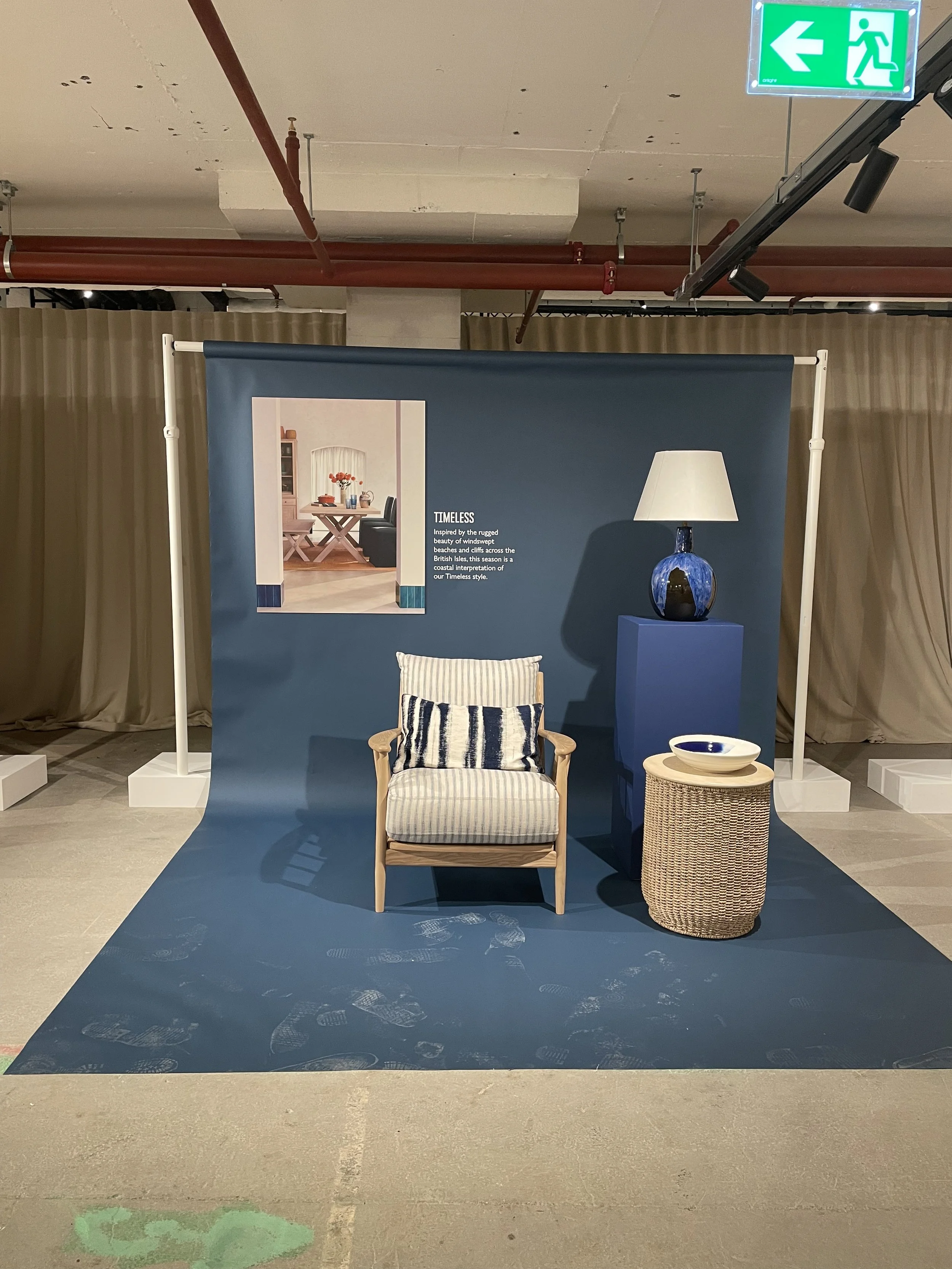 Display of furniture and home decor in a showroom setting with a blue backdrop, including a framed picture, a wooden armchair with striped cushions, a lamp on a blue pedestal, and a wicker side table with a bowl on top, with an emergency exit sign vi