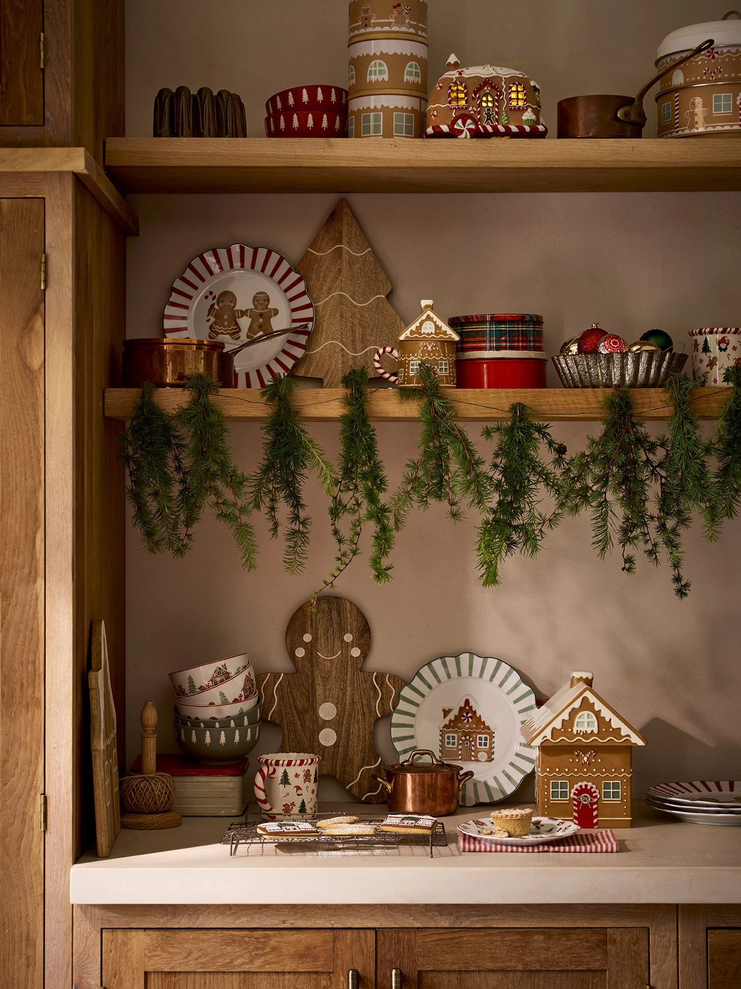 Christmas-themed kitchen shelf decorated with gingerbread cookies, gingerbread house, holiday plates, cups, and greenery garland.