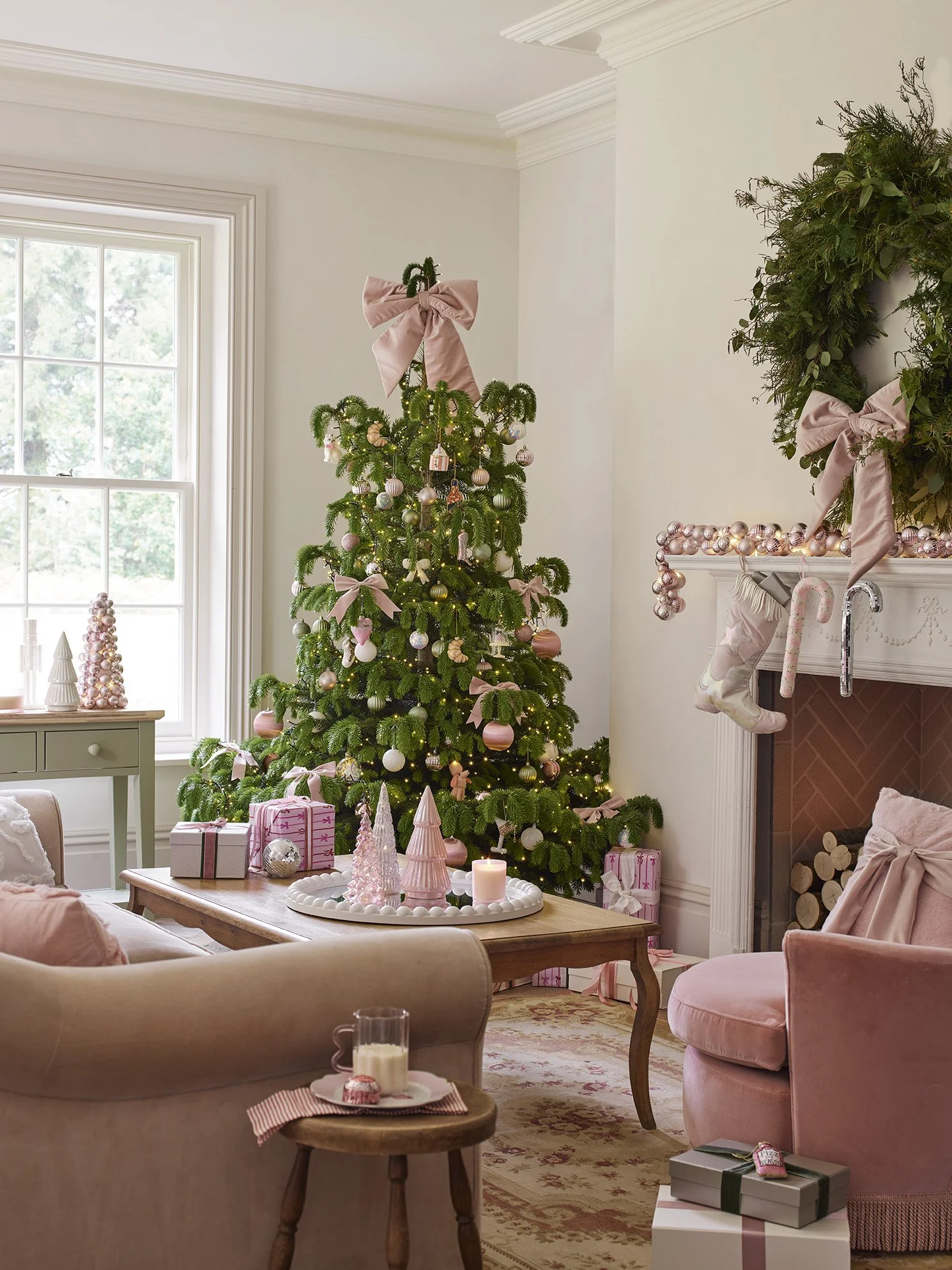 A decorated Christmas tree with pink and white ornaments, bows, and lights in a cozy living room with a fireplace, wrapped gifts, and holiday decorations.