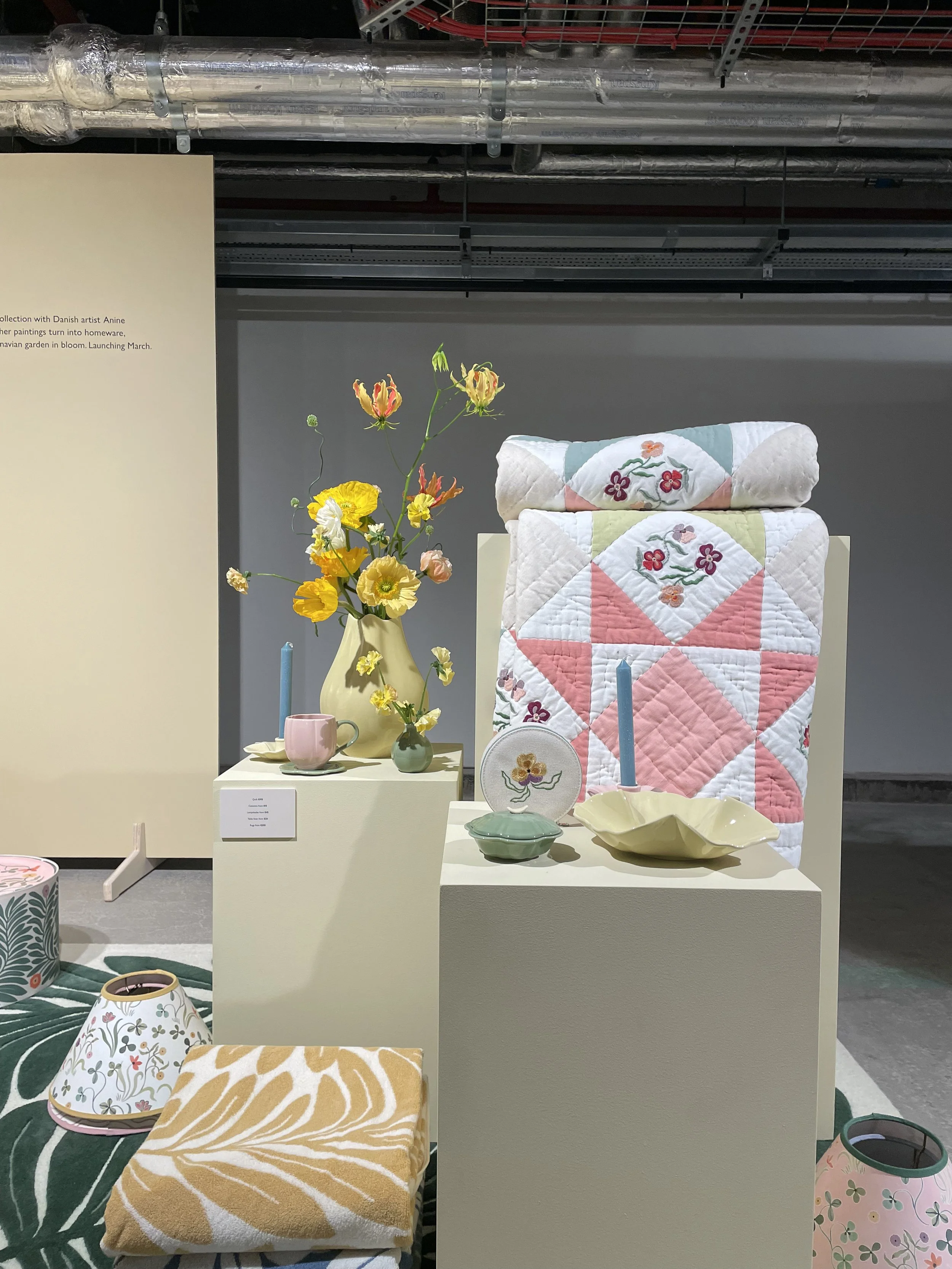 Display of pastel-colored embroidered patchwork quilts, ceramic vases with yellow and pink flowers, decorative candles, a ceramic cup, and a fabric cushion with a leaf pattern on a table in an art exhibit.