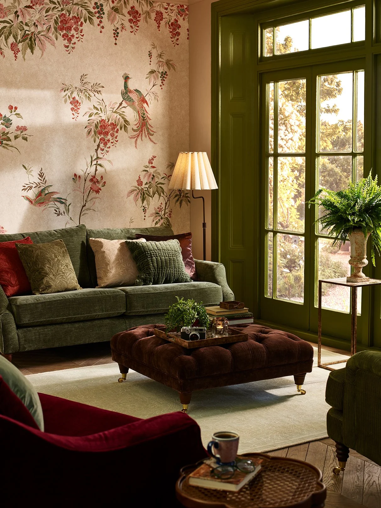 A cozy living room with a green window, a green sofa with patterned pillows, a brown tufted ottoman with plants and a camera, an armchair with a coffee cup, and a wallpaper featuring a bird and floral design.