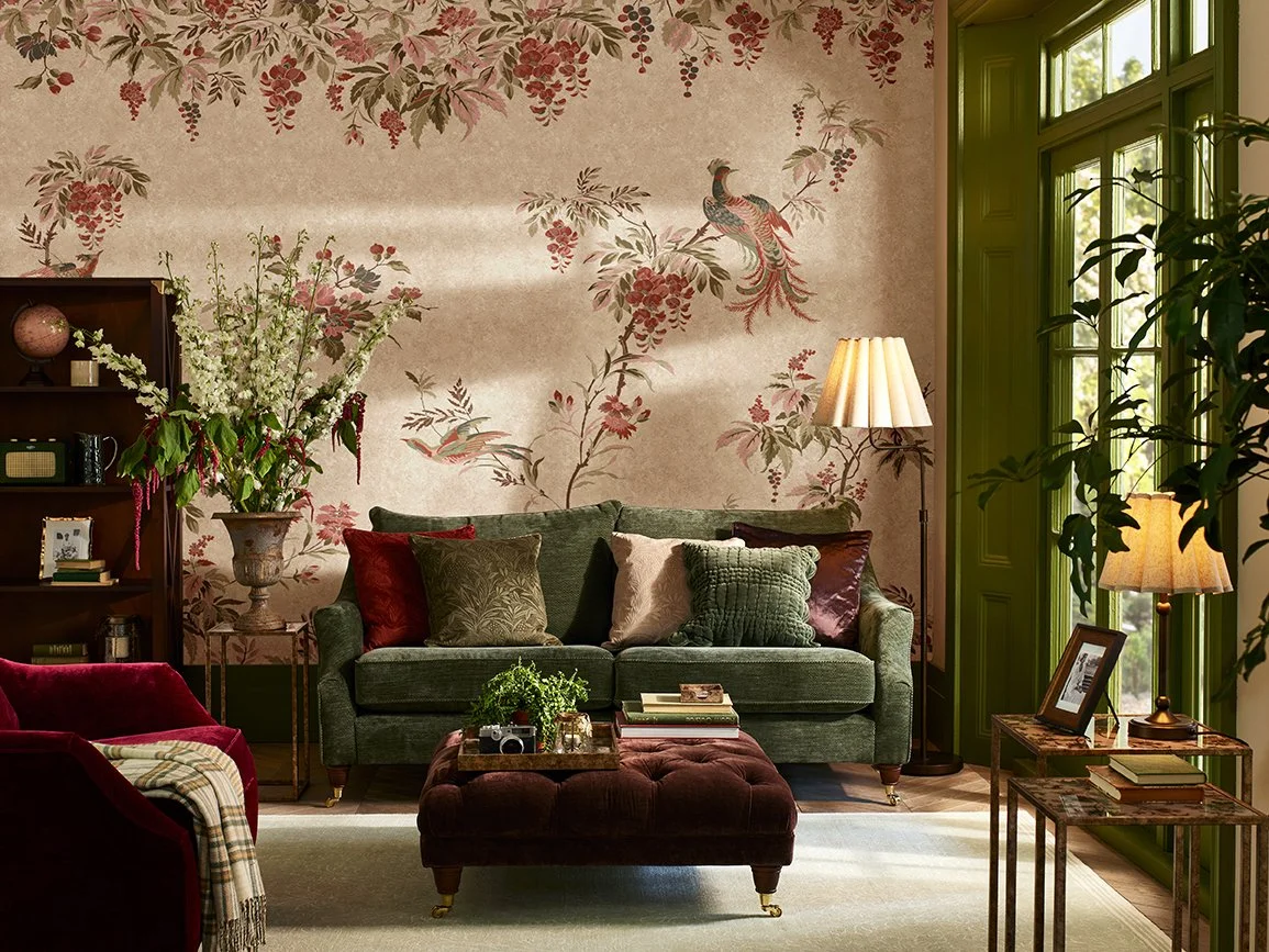 Living room with green sofa, red armchair with plaid blanket, and ottoman, floral wallpaper with peacock design, floor lamps, side tables with books and framed photo, floral arrangement on side table, and large windows with green trim.