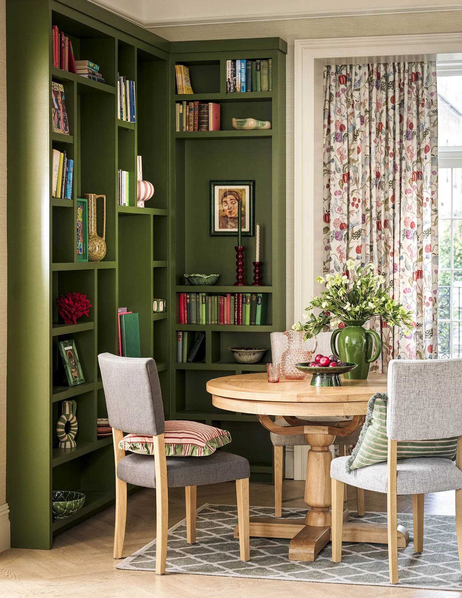 A living room corner with a green built-in bookshelf filled with books and decorative objects, a round wooden dining table with a large green vase containing white flowers, and three chairs around the table. A window with floral curtains is visible i