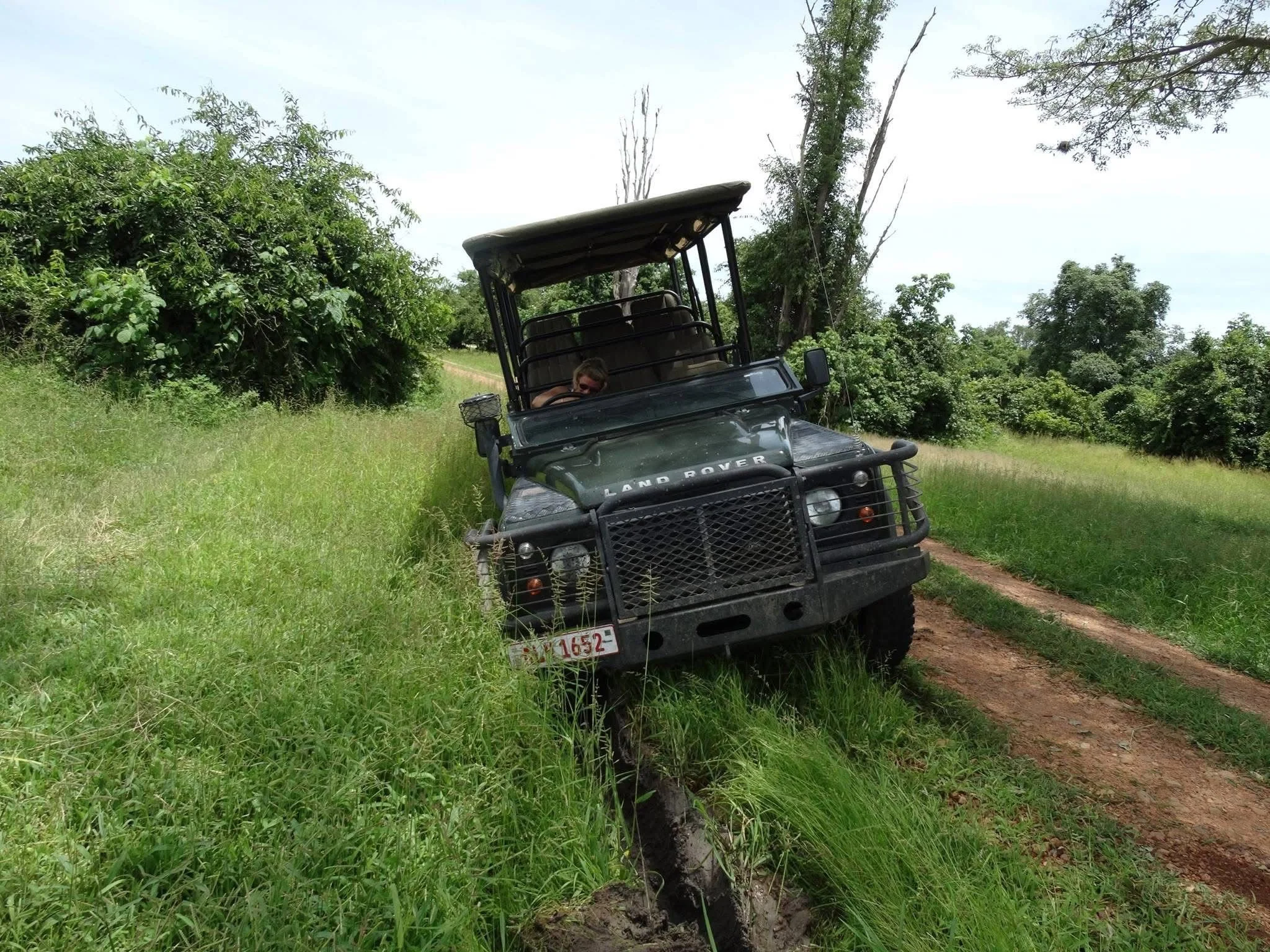 A black Land Rover vehicle parked on a grassy hillside next to a dirt trail amid trees and greenery in a safari lodge in Zambia.