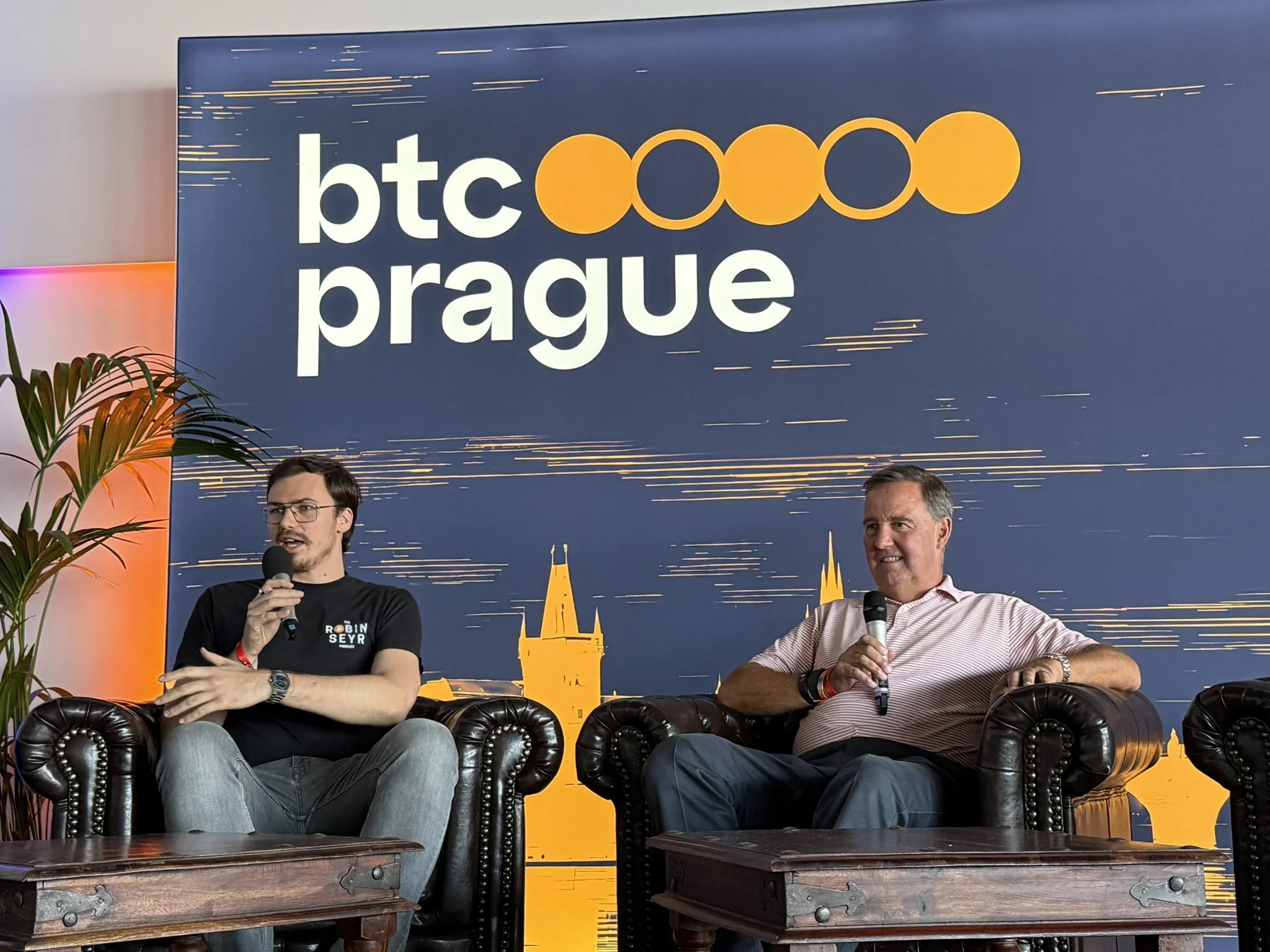 Two men are seated on black leather armchairs on stage at BTC Prague event, speaking into microphones. Behind them is a large blue banner with the BTC Prague logo and an orange and white skyline silhouette.