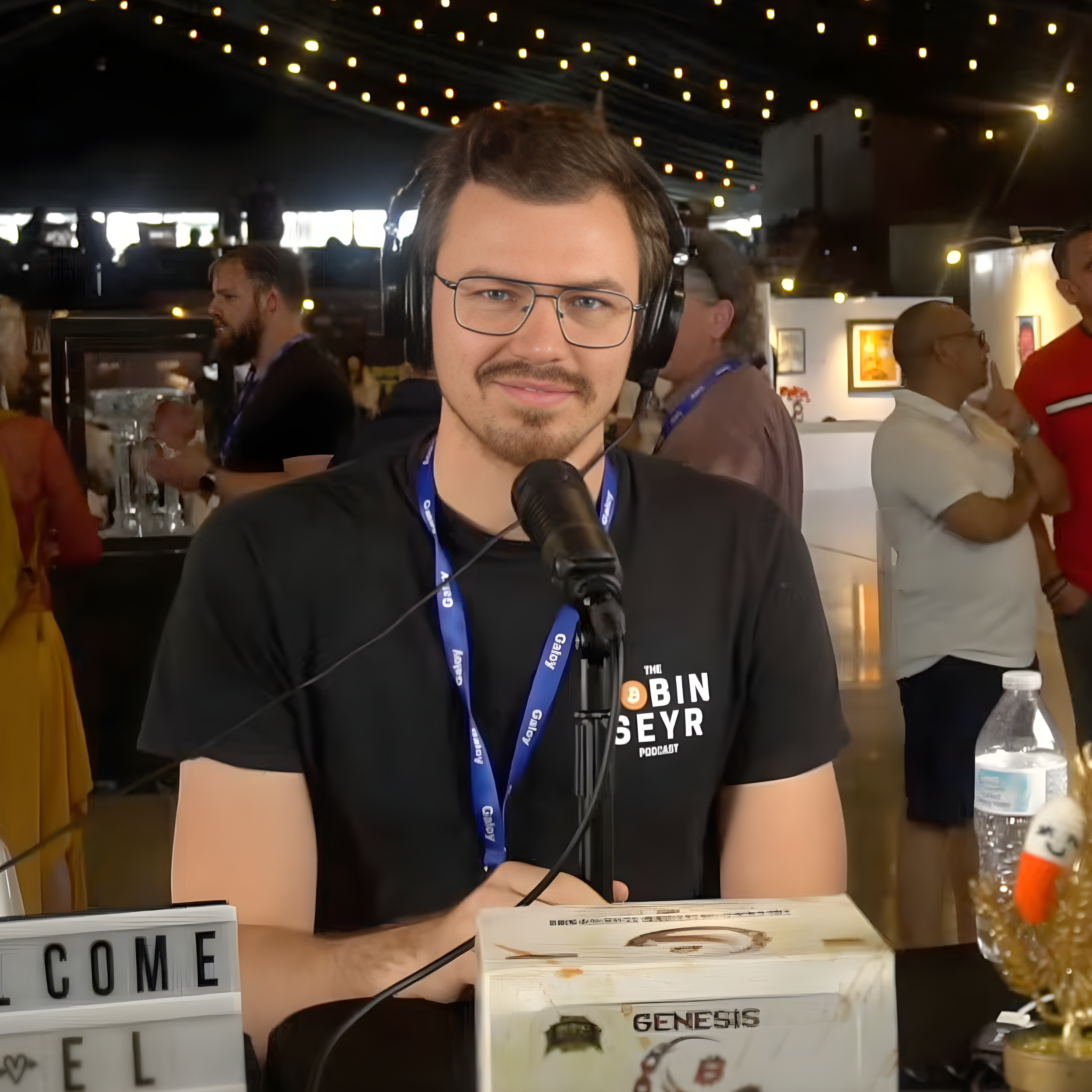 Man with glasses and headphones speaking into a microphone at an event with other people in the background.
