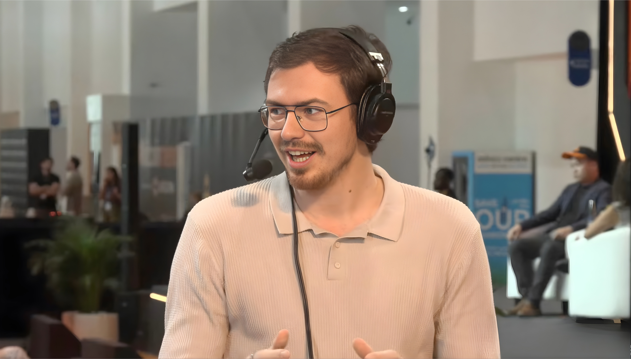 A man with glasses and a beard wearing a beige shirt and a headset with a microphone, smiling and talking, in an indoor public space with people sitting and standing in the background.