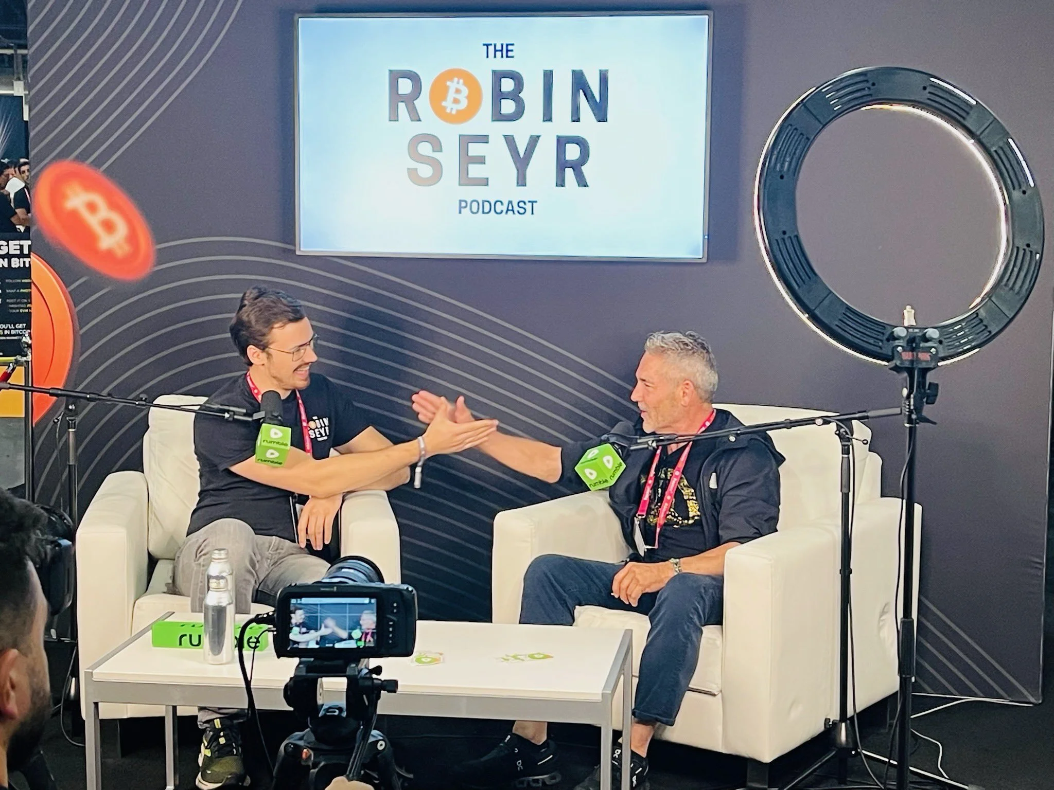 Two men sitting on white sofas engaging in a handshake during a podcast recording at the Robin Seyr Podcast, with a large screen behind them displaying the podcast title, and a camera in the foreground capturing the scene.