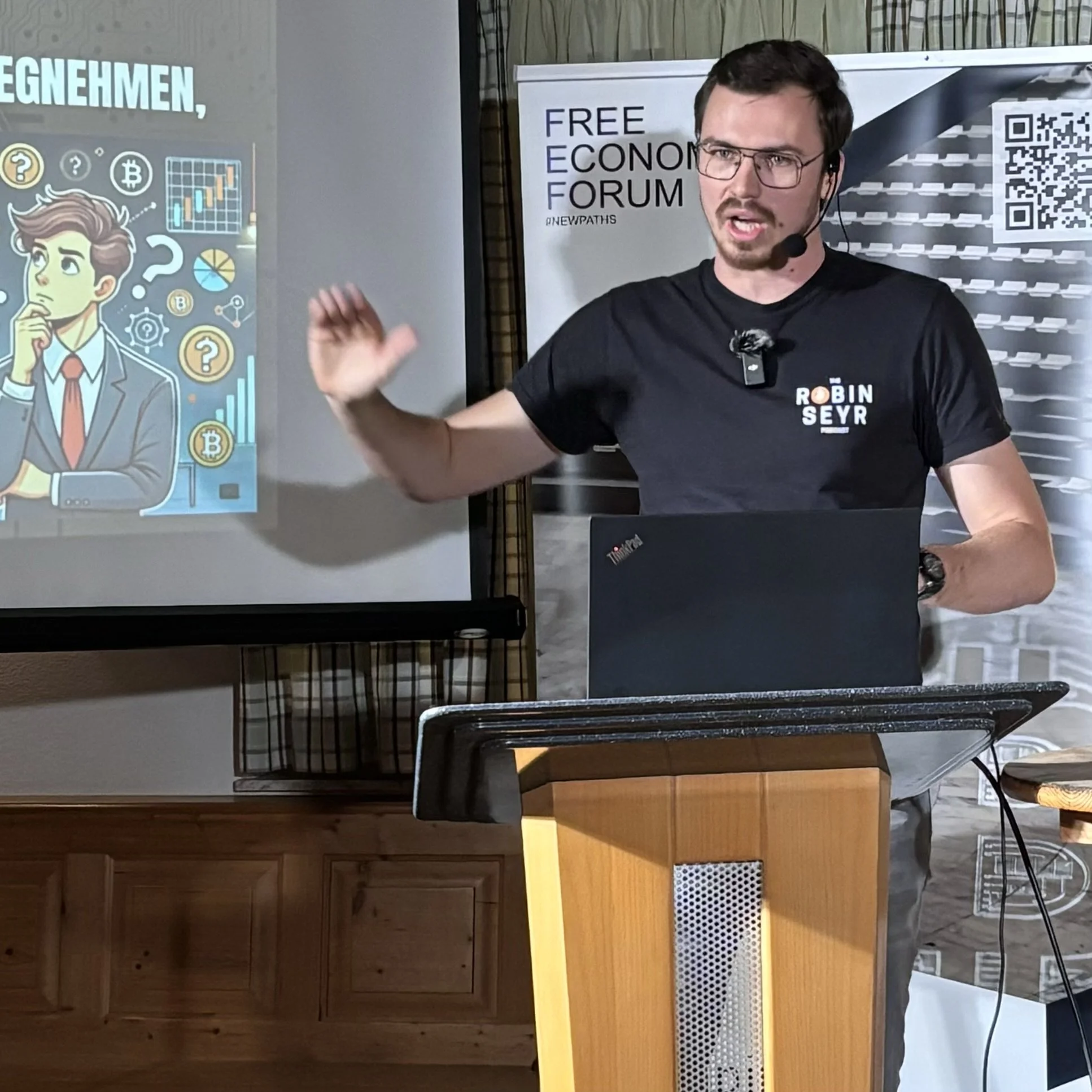 A man with glasses and a black t-shirt, who has a microphone headset, is giving a presentation at a podium. Behind him, there is a projection screen showing a cartoon of a thinking man with financial charts and Bitcoin symbols. To the right, there is a banner with the text 'FREE ECONOMY FORUM' and a QR code.