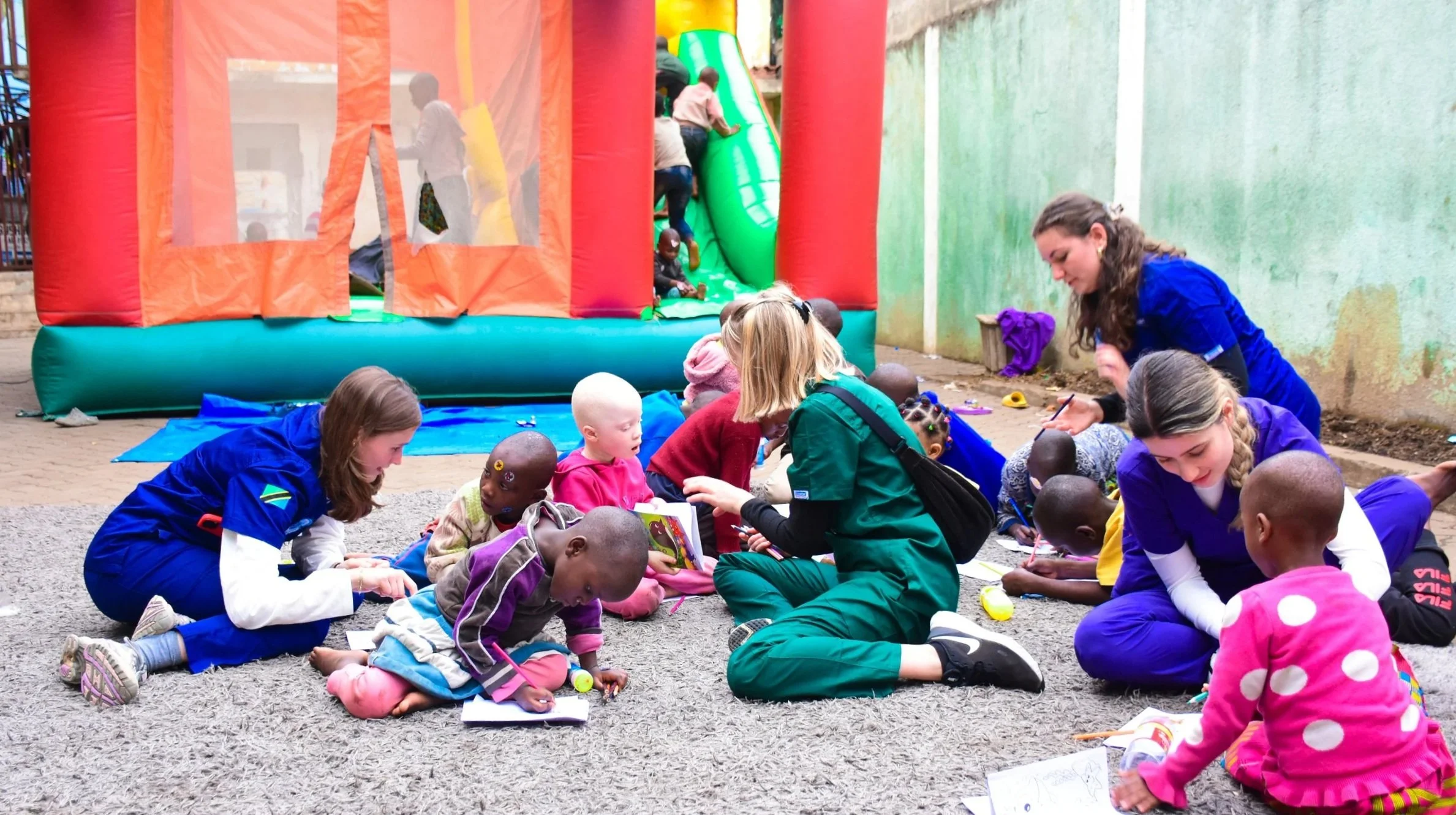 Children and adults sitting on the ground, coloring and drawing, with a colorful inflatable bounce house in the background.