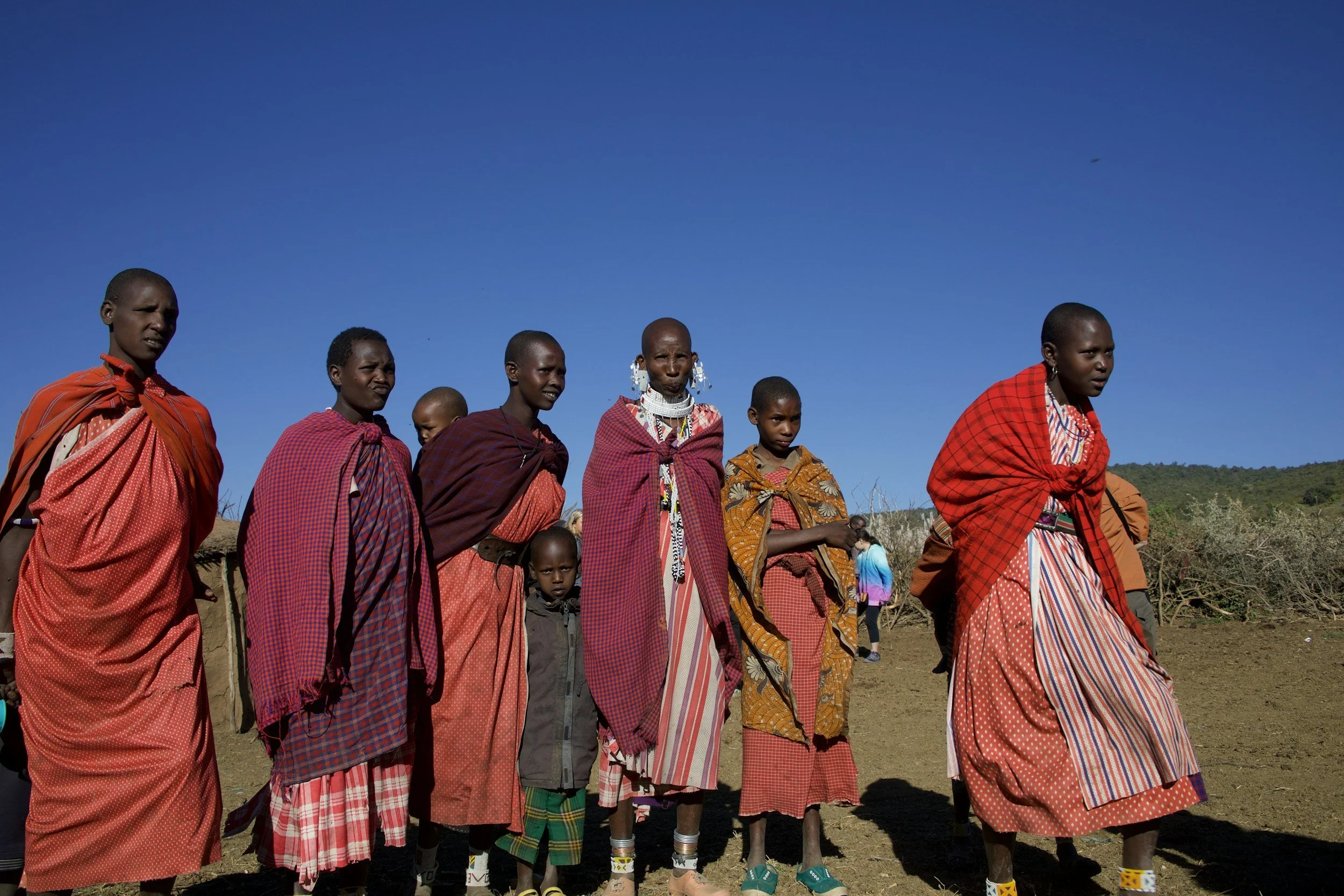 Group of Maasai women and children standing outdoors against a clear blue sky, dressed in traditional red and patterned shukas.