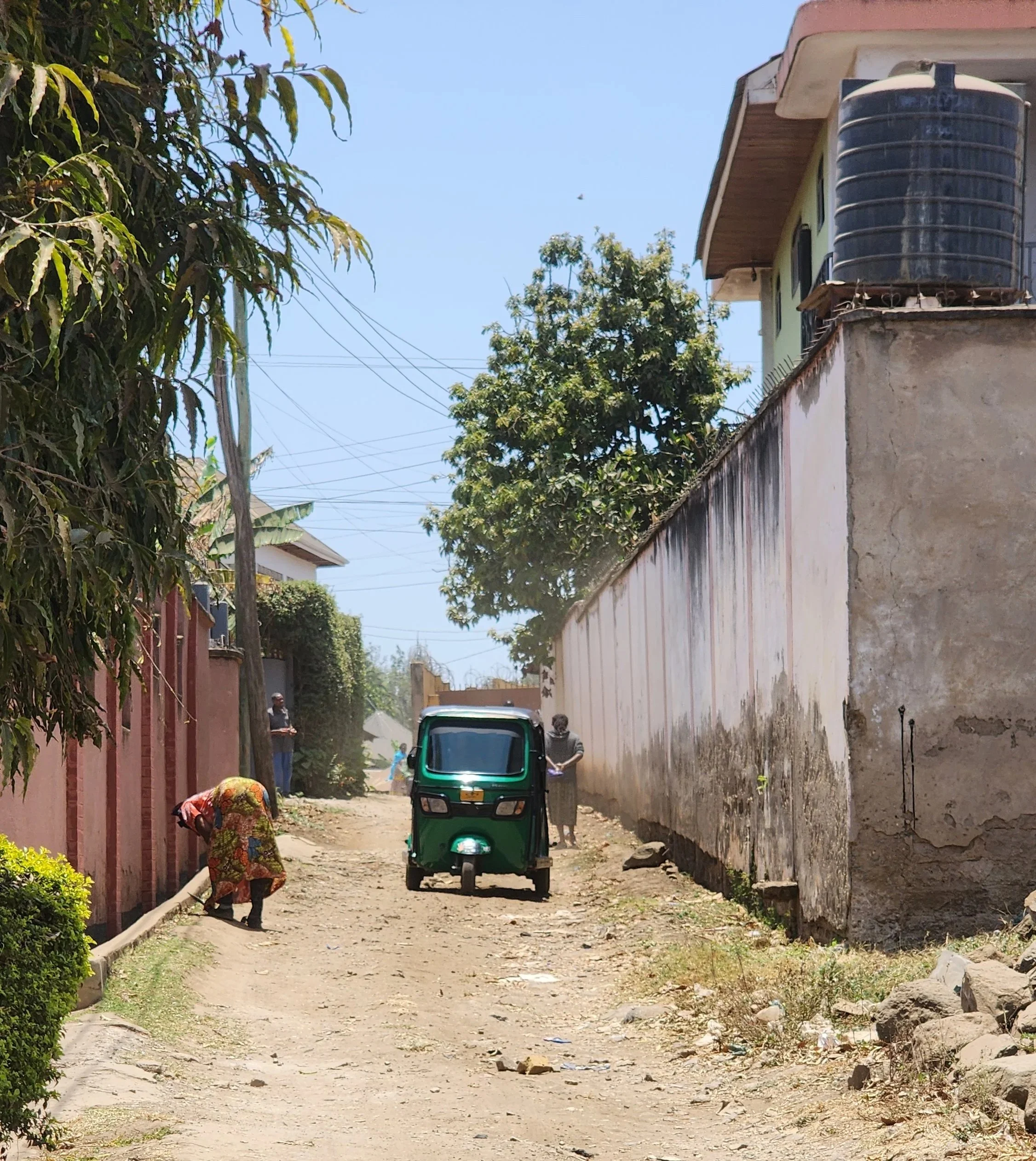 A dirt alleyway with a green auto rickshaw and several people, including a woman bending down and another person standing near the auto, flanked by buildings and trees under a clear blue sky.