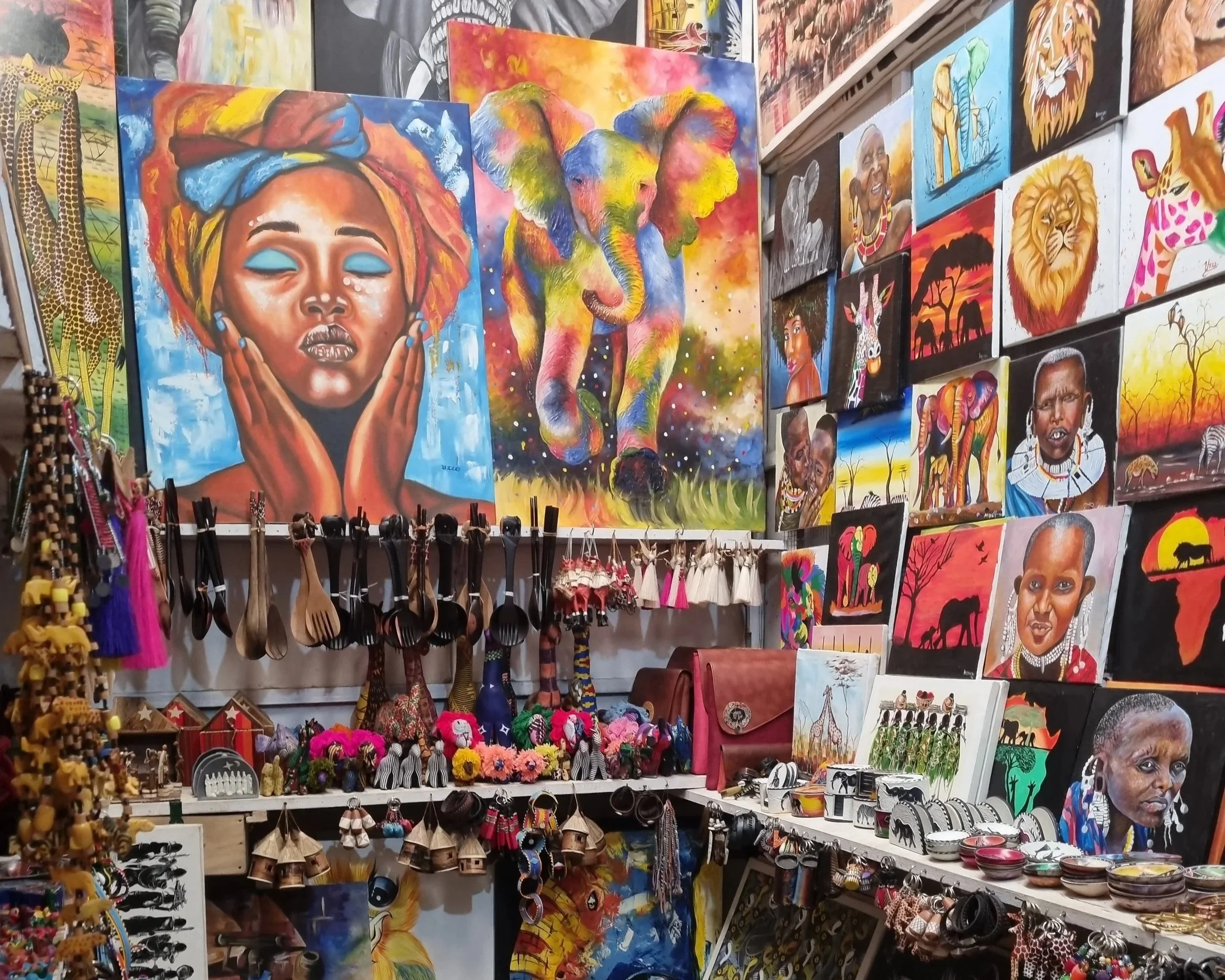 Colorful art gallery wall featuring African-inspired paintings of people, animals, and landscapes, along with handmade jewelry and accessories displayed below in Arusha, Tanzania.