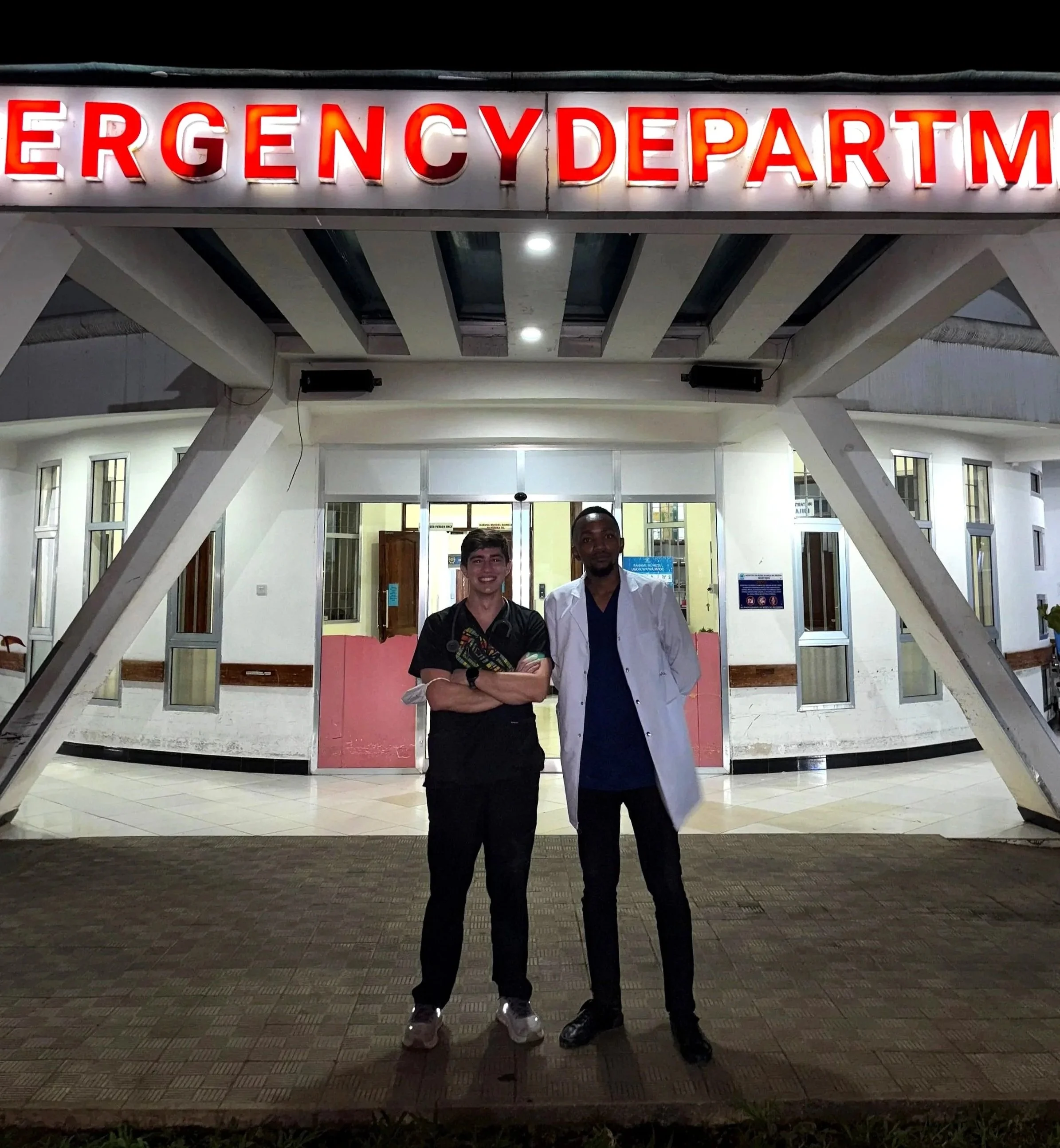 Two young men standing outside an emergency department entrance at night, smiling and posing for the photo.
