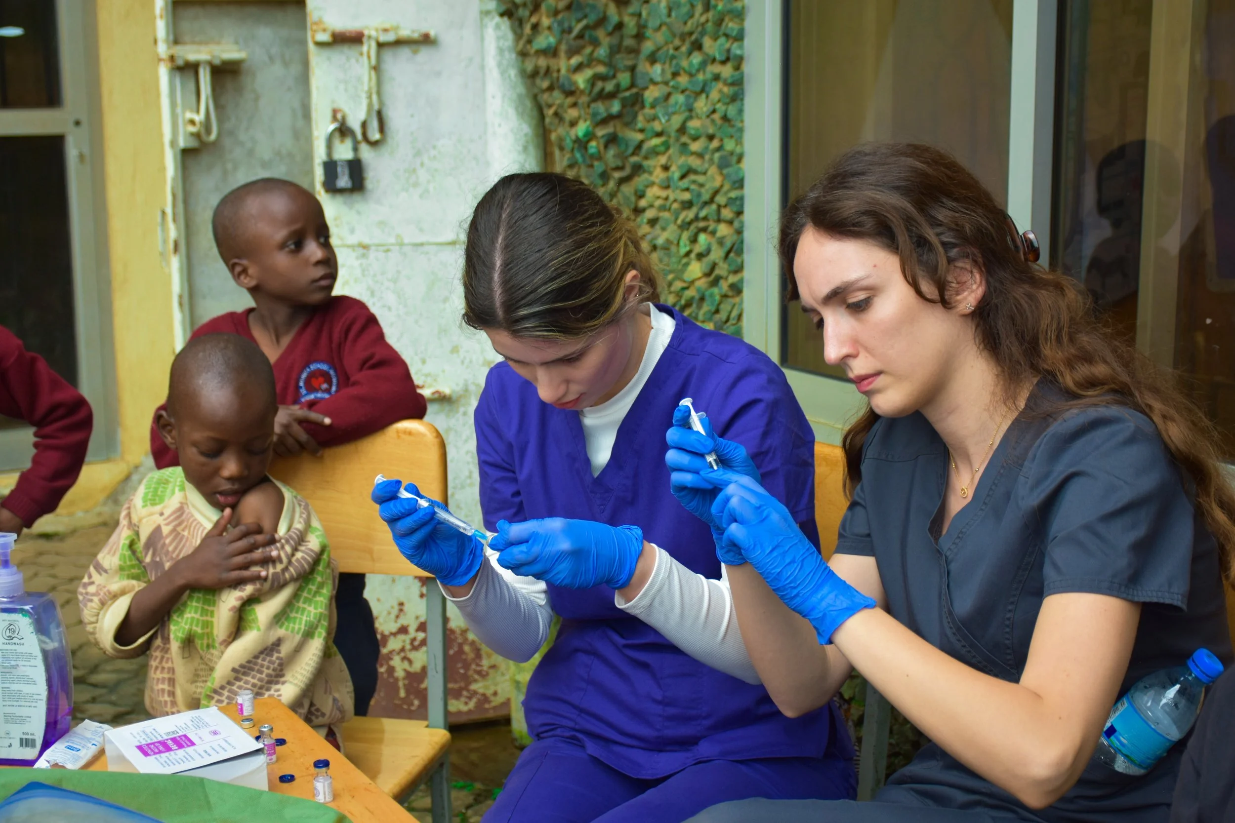 Two female healthcare doctor volunteers with blue gloves preparing syringes while children watch in an outdoor setting, preparing to vaccinate orphaned children against disease.