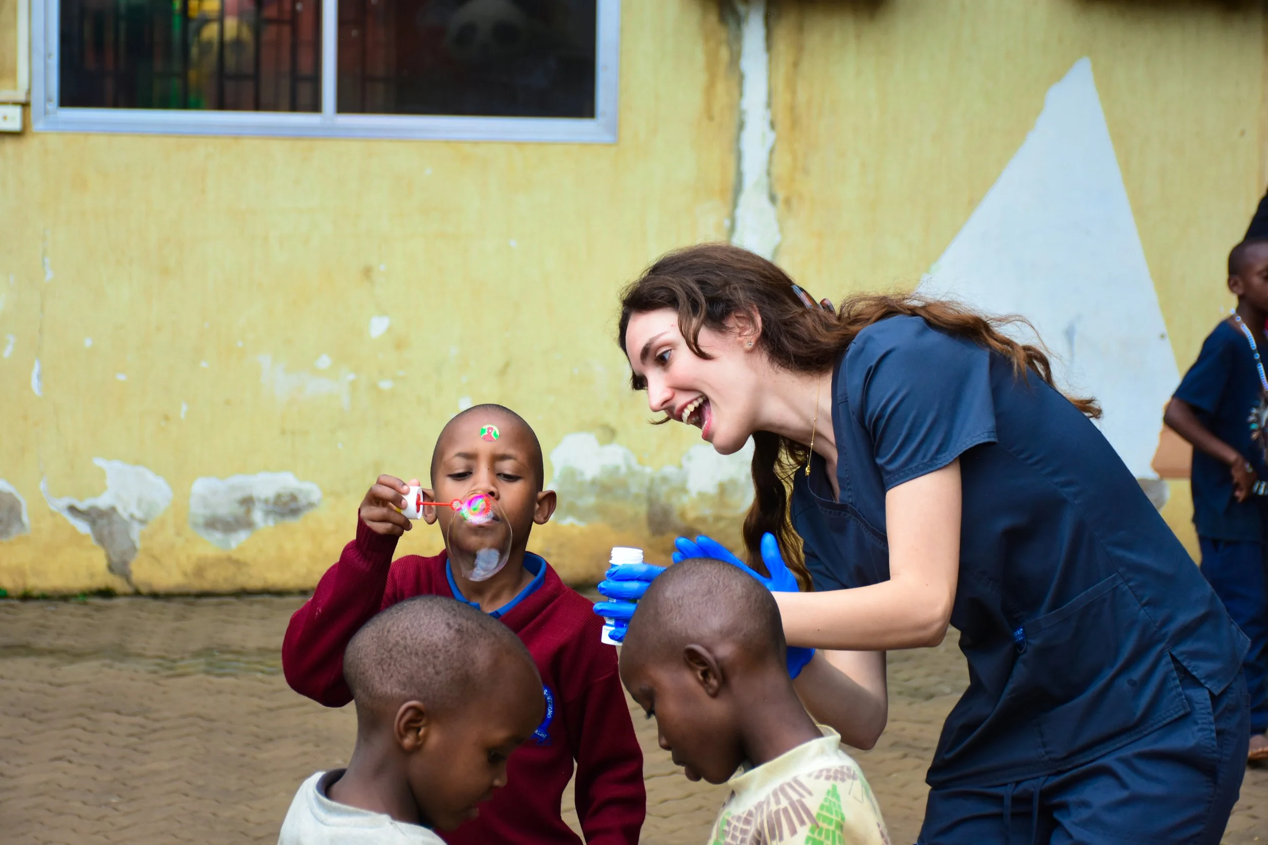 A female dental volunteer with long brown hair, wearing a blue medical scrubs and blue gloves, is helping orphaned children blow bubbles outside in an orphanage, with a yellow wall with peeling paint in the background.