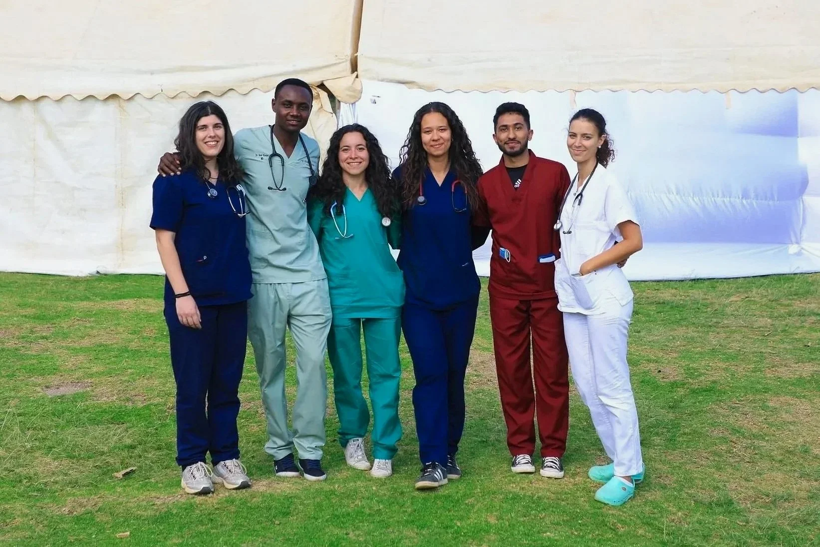 Group of six diverse medical professionals in uniforms standing outdoors on grass in front of a white tent, smiling at the camera.