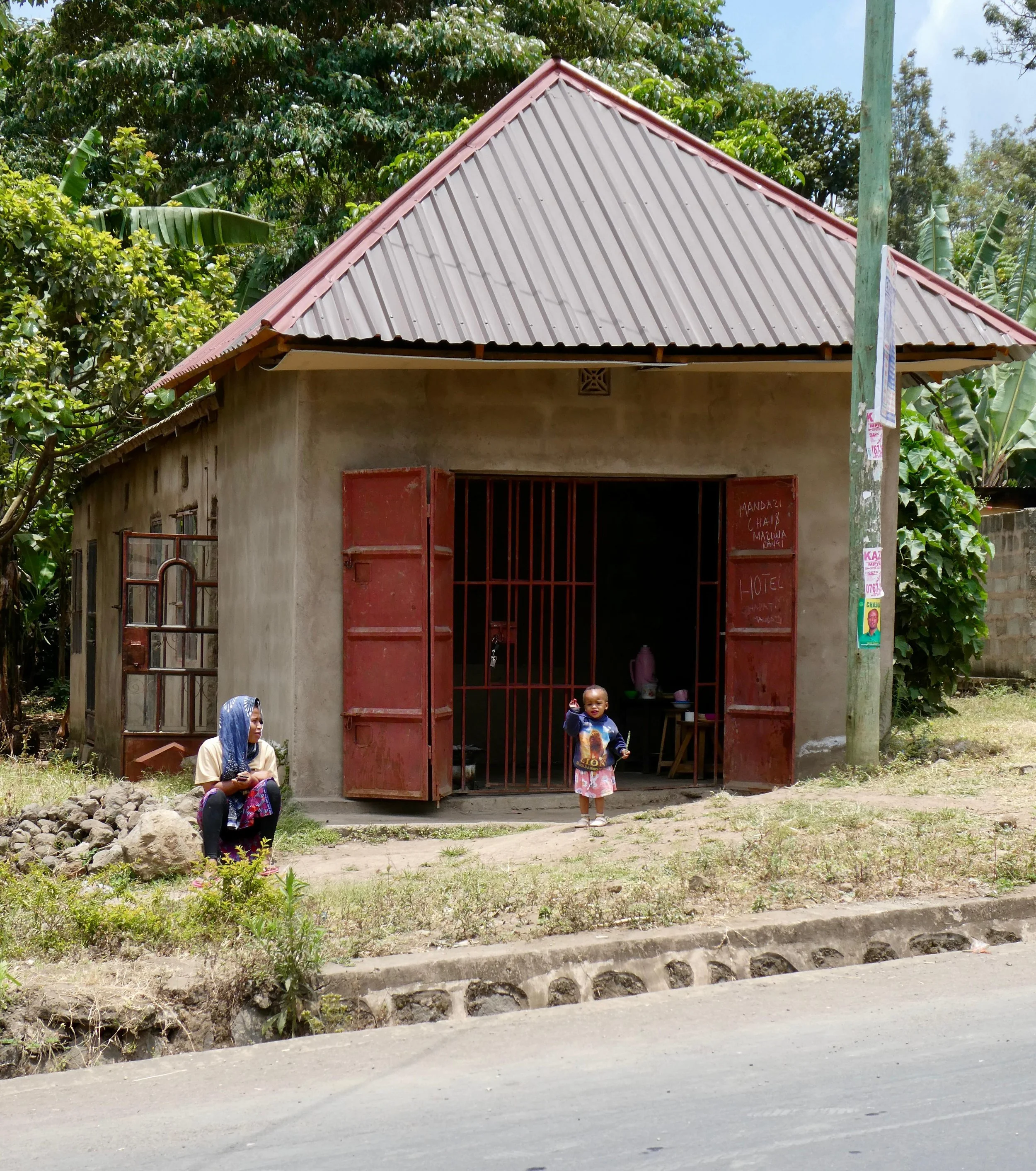 A small concrete building with open rusty metal doors, situated beside a dirt road and surrounded by green trees. Two children are outside; one is sitting on rocks near the house, and the other, a young girl, is standing in front of the door waving. 